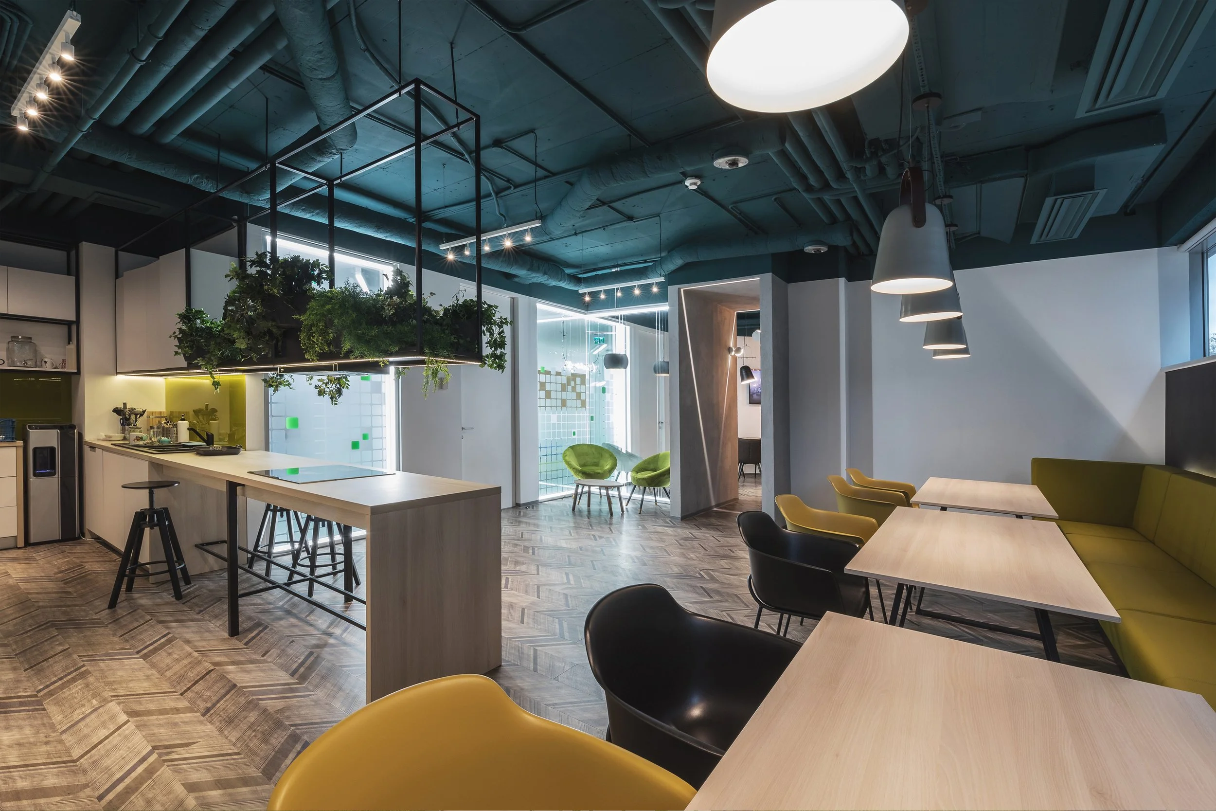 Modern office or café space with wooden tables and chairs, green seating area, and overhead lighting fixtures.