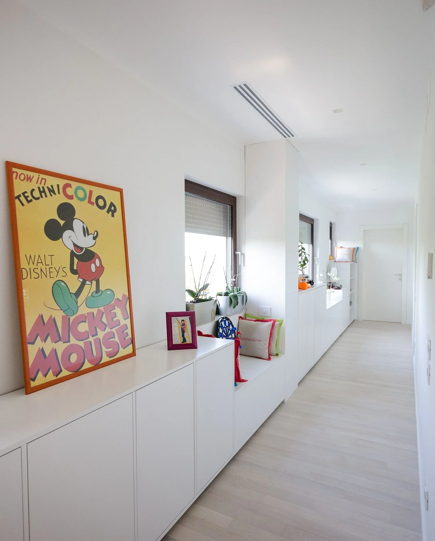A bright, minimalist hallway with white walls and light wooden flooring. There are two small windows with plants on the window sill, several cushions with colorful borders, and framed pictures on a long white cabinet. A whimsical Mickey Mouse poster decorates the wall.