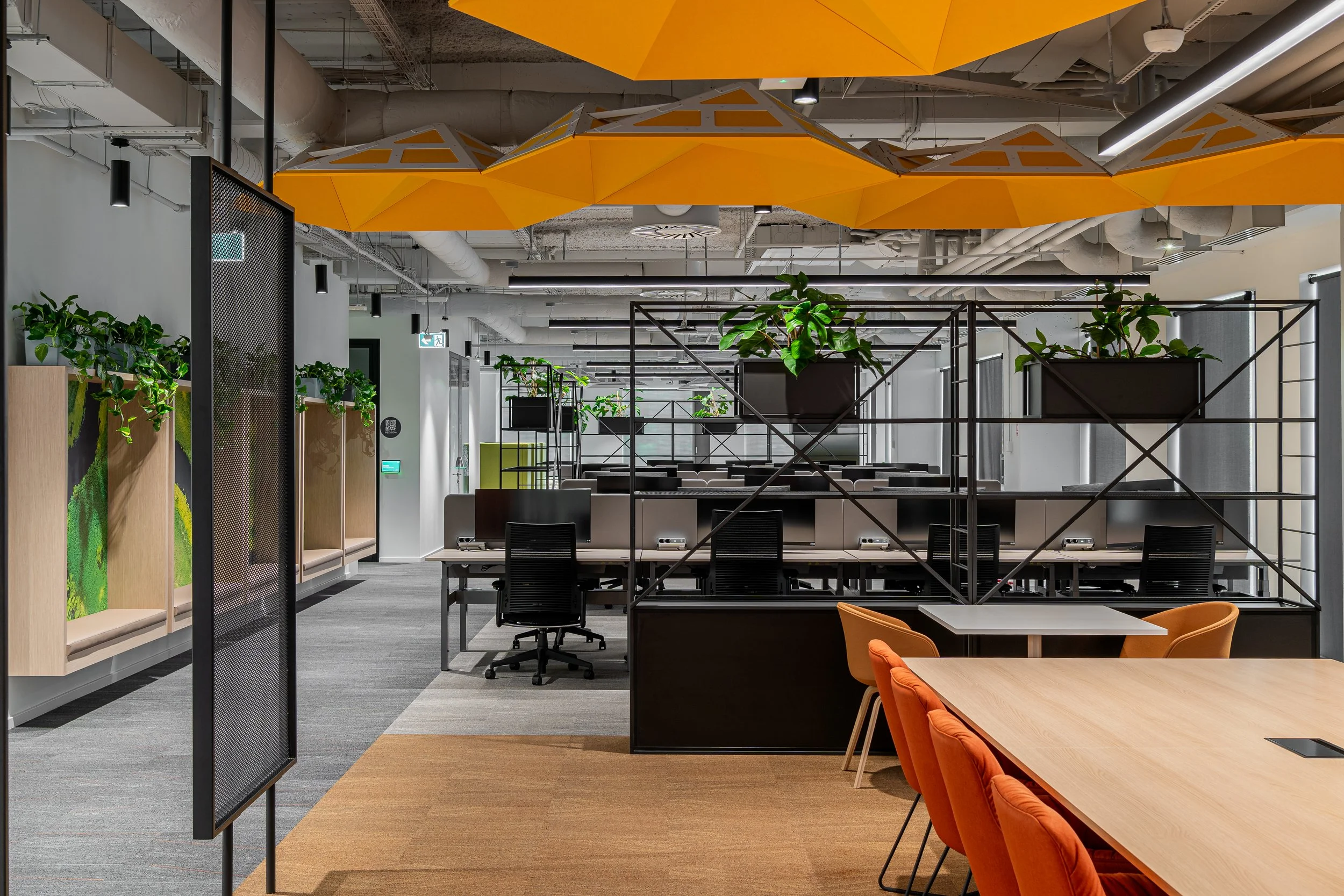 Modern office workspace with orange and black chairs, wooden and white tables, green plants, black metal shelves, and orange ceiling panels.