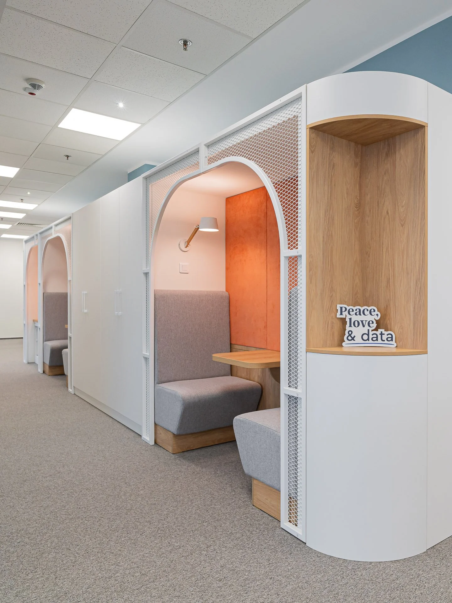 Modern office or lounge area with white booths that have upholstered seating, small built-in tables, and decorative lighting, with a sign reading "Peace, love & data" on one of the wooden panels.