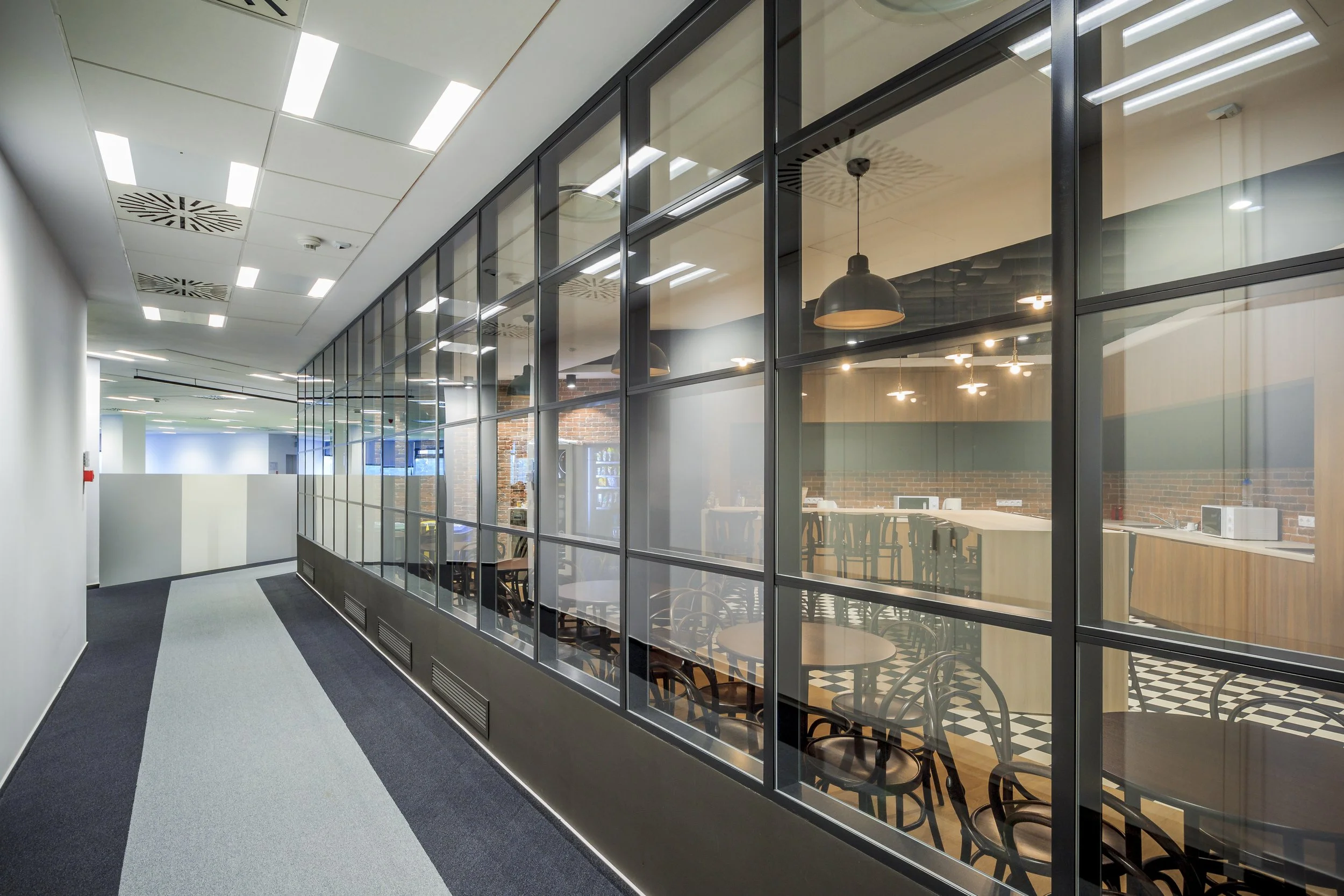 Empty office corridor with a glass partition, conference room with tables and chairs, modern lighting, and a brick wall inside.