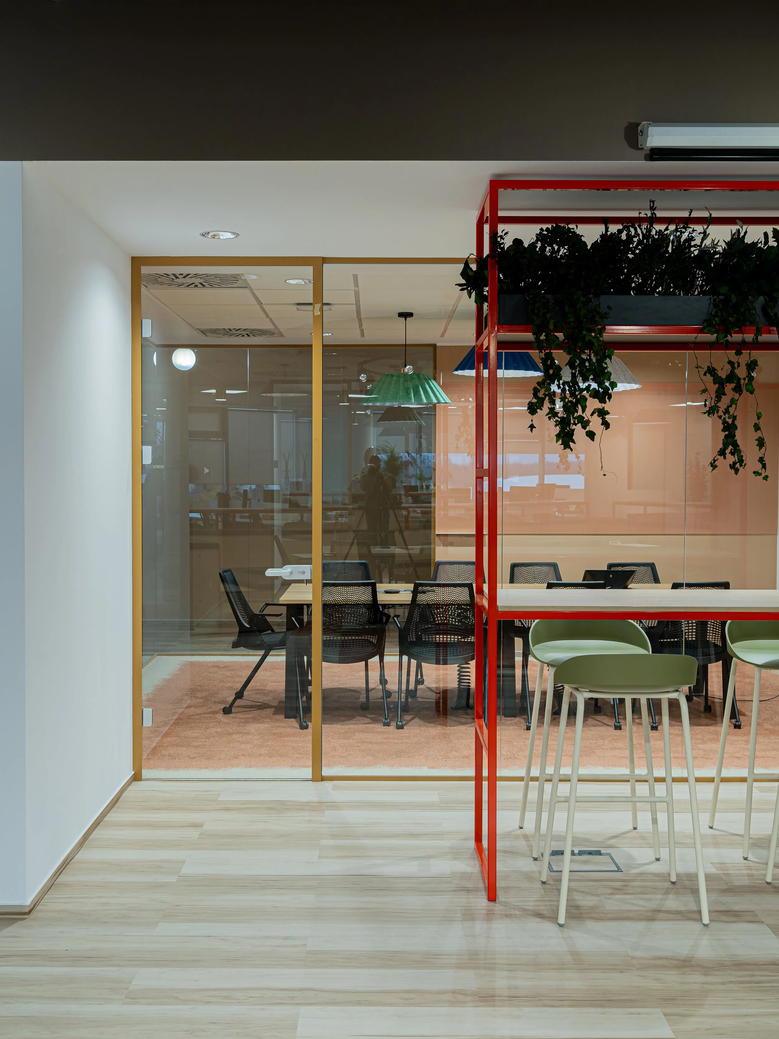 Interior view of an office space with a glass-walled meeting room, bar-height table with green chairs, and a red metal shelf with greenery.