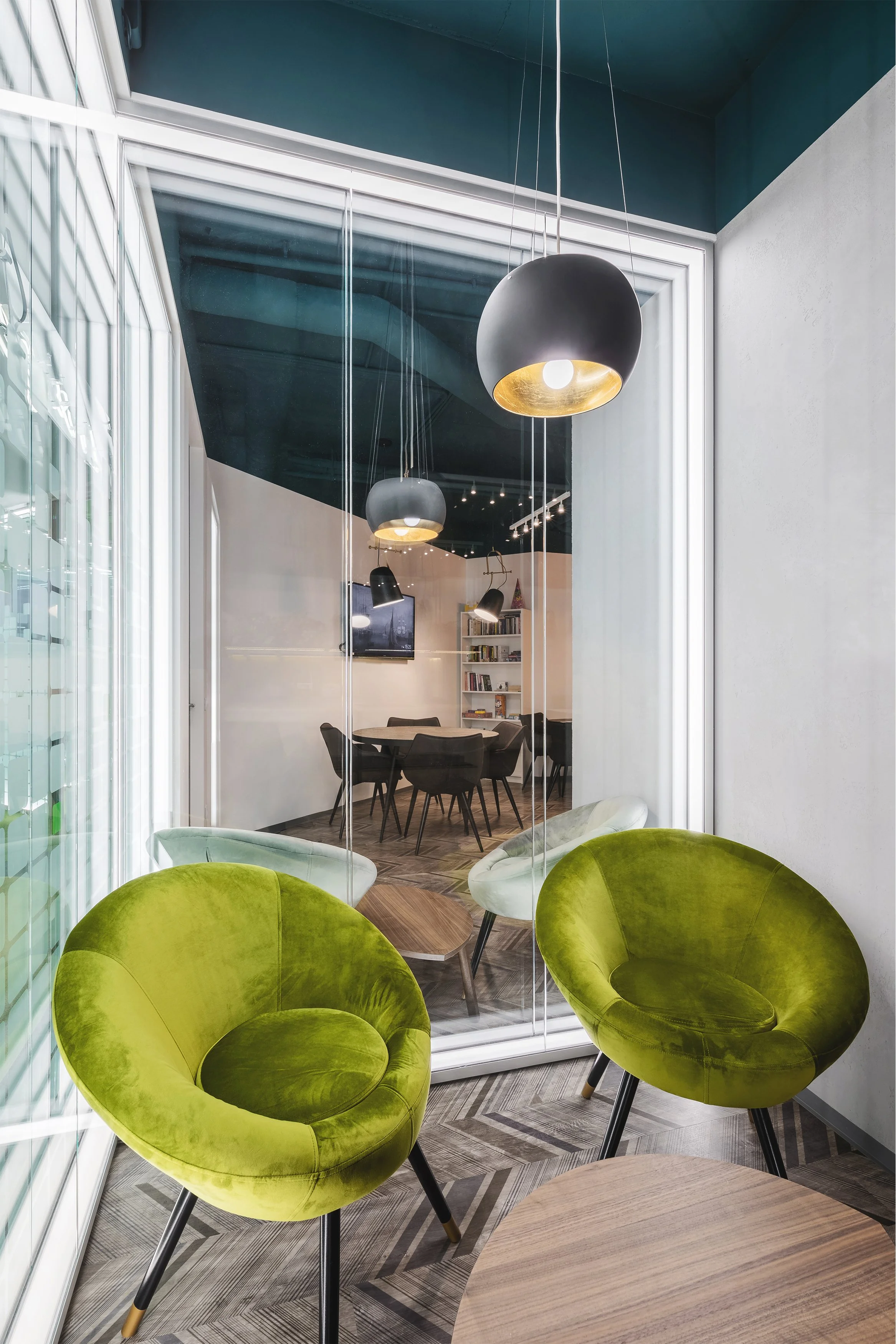 Modern office space with bright green velvet chairs, white walls, and a glass wall overlooking a conference room with a round table and black chairs, illuminated by black and gold pendant lights.