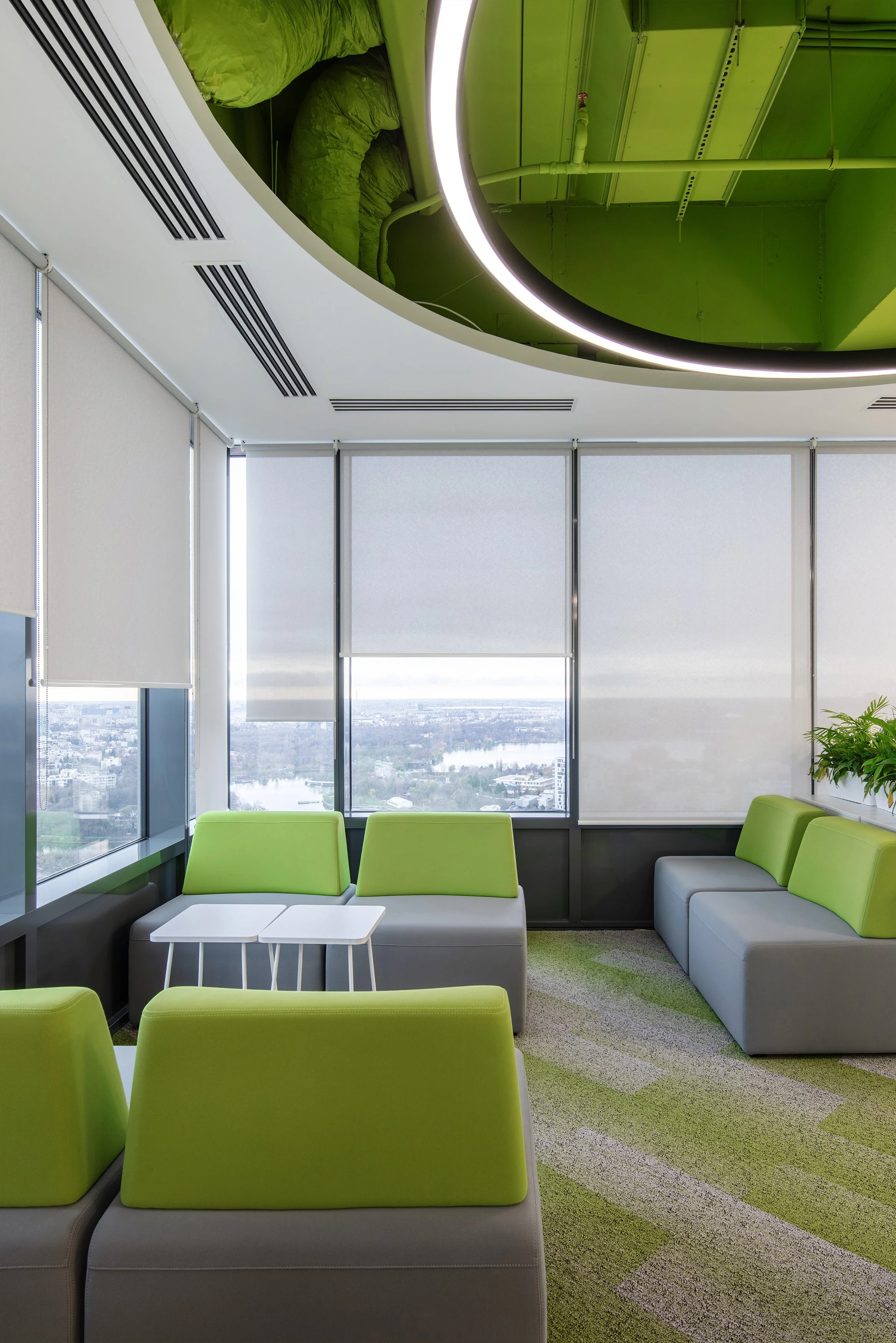 Modern office lounge with lime green and gray seating, white tables, large windows with blinds, overlooking a cityscape, with a green ceiling feature and round lighting.