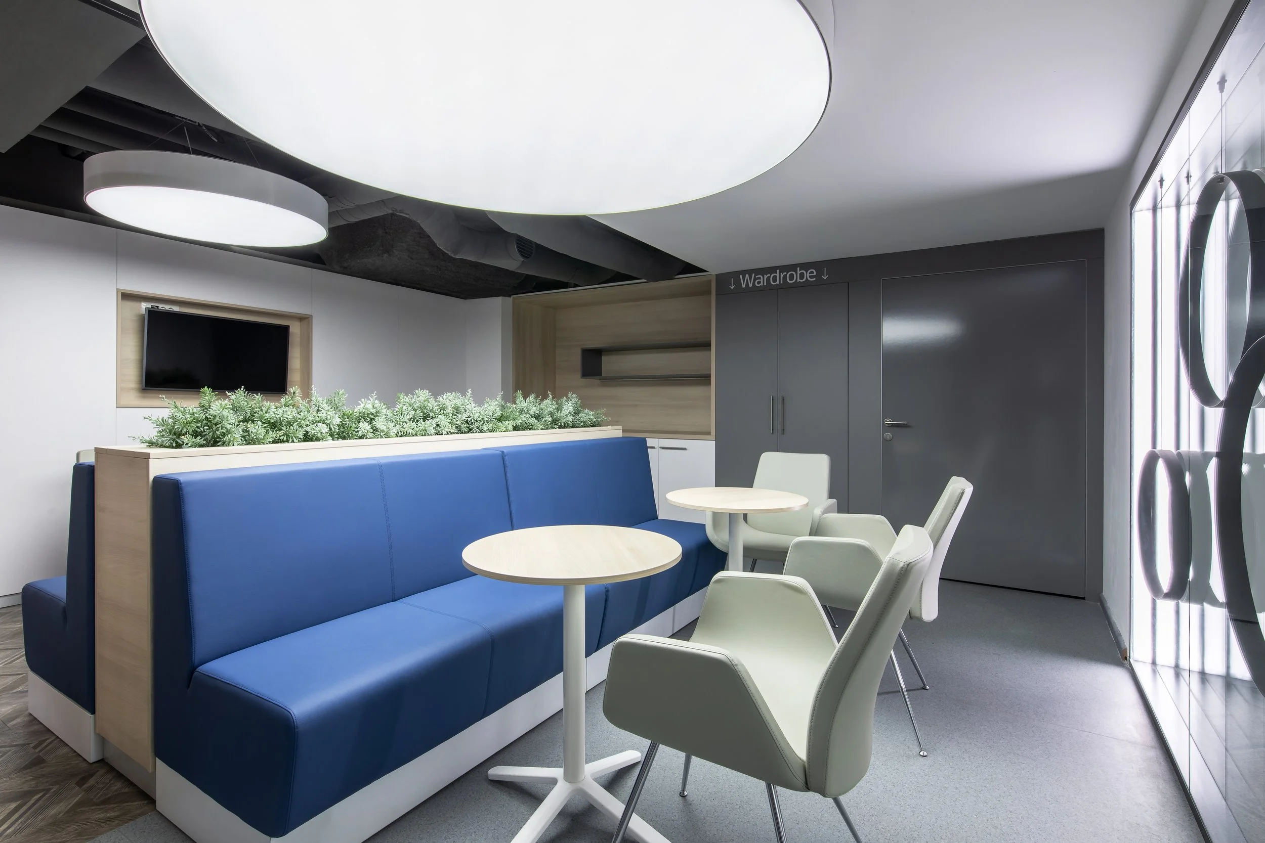 A modern lounge area with blue cushioned bench seating, white chairs, and small round tables, featuring a flat-screen TV, indoor plants, and a sign indicating a wardrobe.