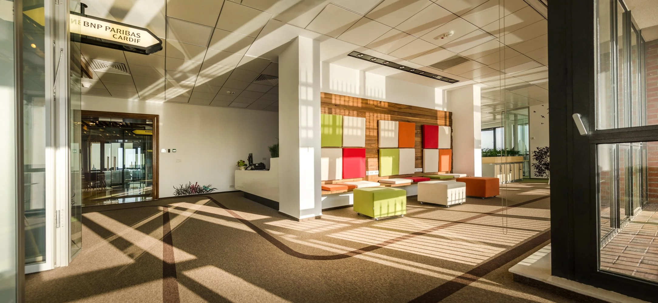 Modern office lobby with colorful seating area, natural sunlight, and decorative wall art, leading to conference rooms.