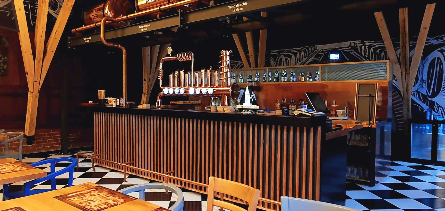 A modern bar with black and white checkered floor tiles, a wooden bar counter with copper pipes and bottles behind it, and a colorful mural in the background.