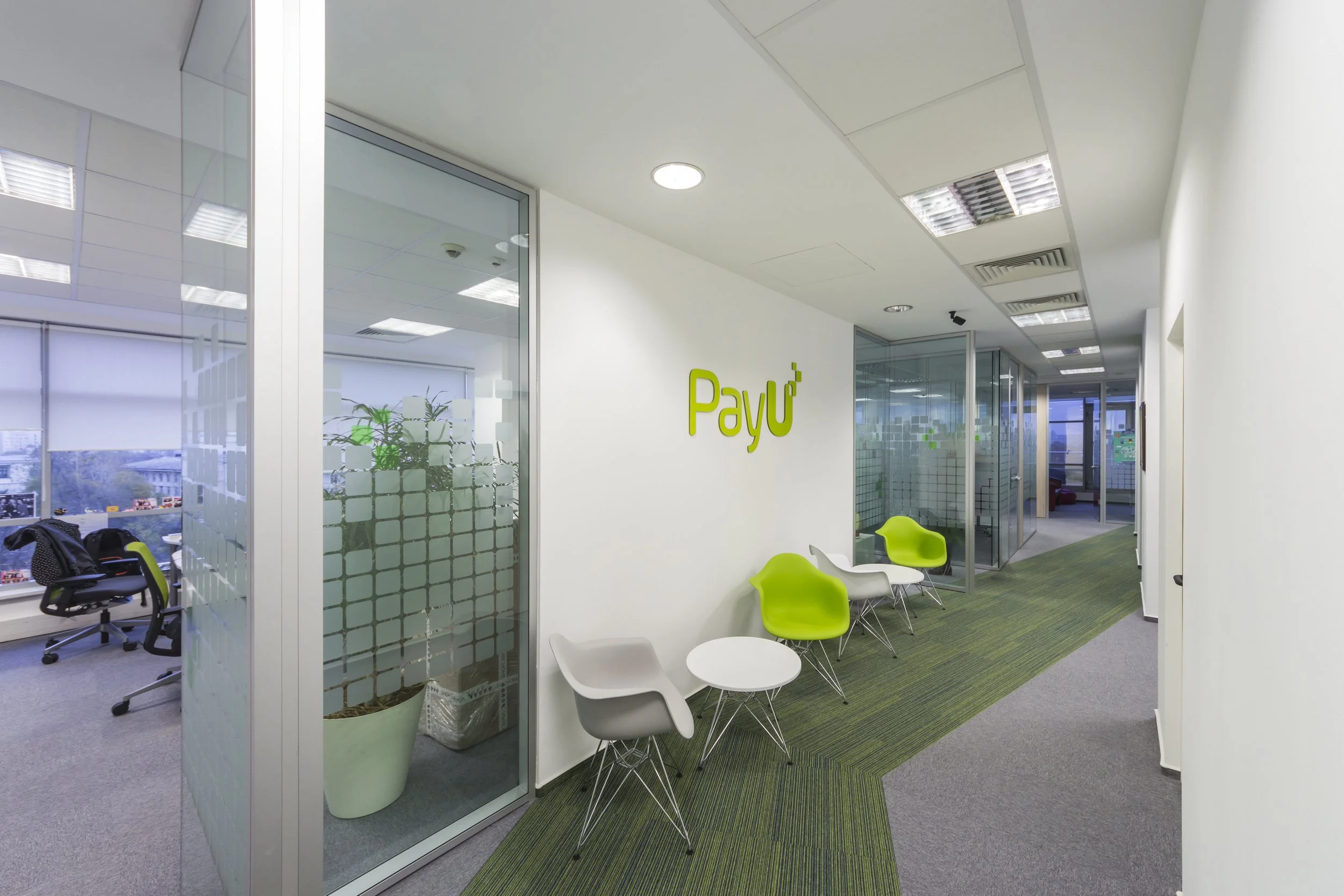 Office corridor with chairs and a wall sign reading 'PayU' decorated with green logo, glass doors leading to offices, and a windowed room visible in the background.