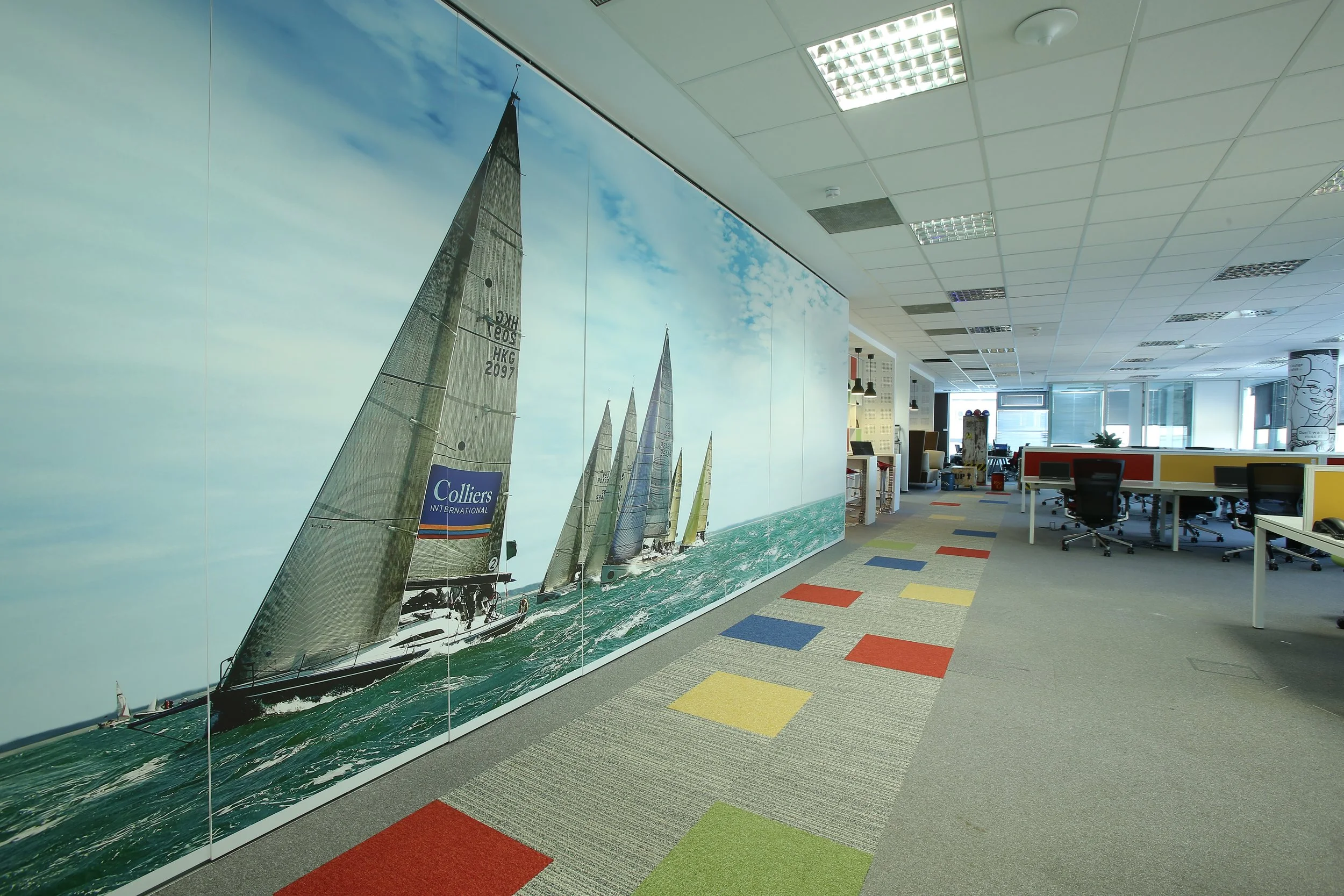 Office space with a large mural of sailing boats on the wall, colorful carpet tiles, workstations, and windows with natural light.