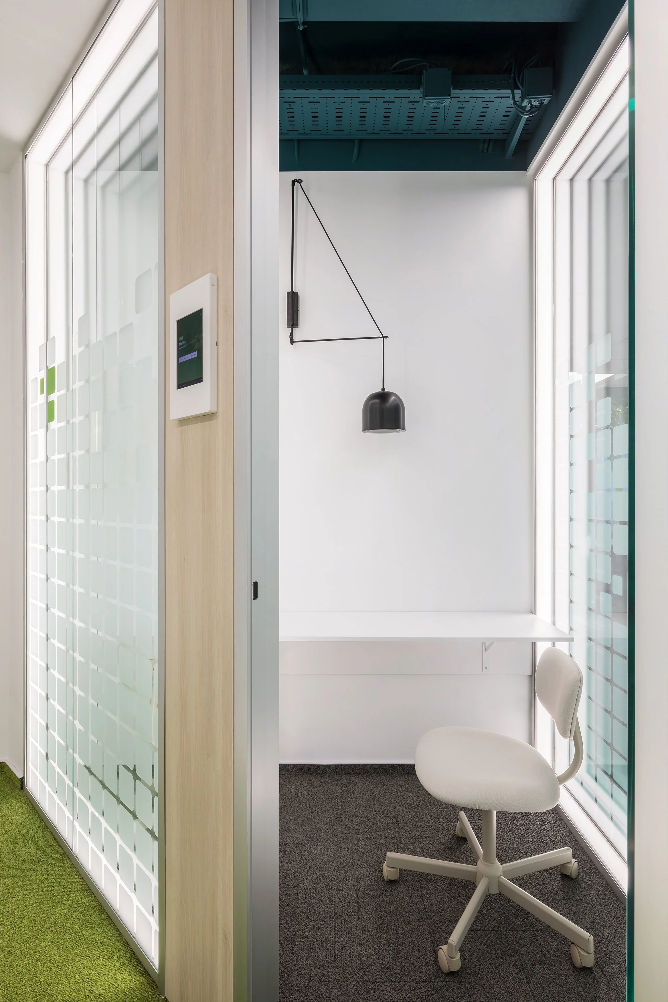 Modern small office cubicle with white walls, a white desk, a white office chair, and a black hanging lamp. Surrounding glass walls with frosted patterns and LED lighting, with a green carpet on one side and dark carpet on the other.