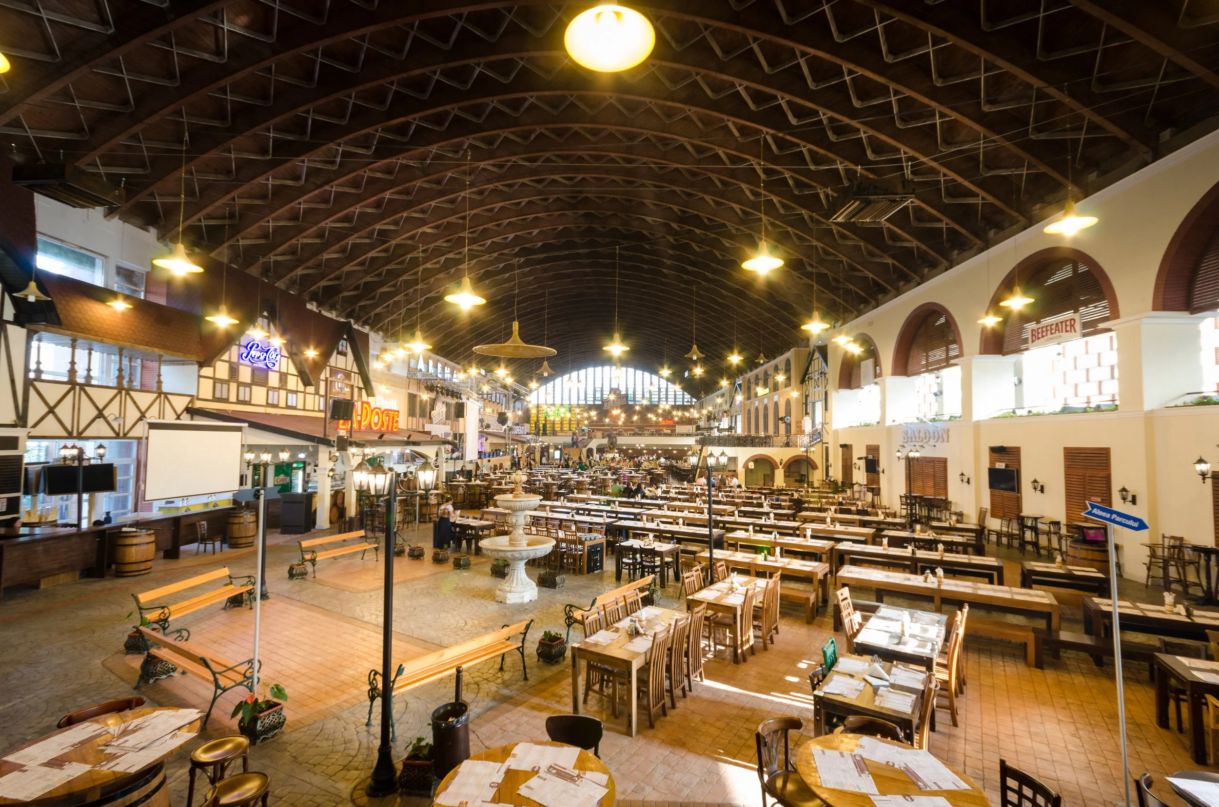 Interior of a large, well-lit restaurant or food court with wooden tables and chairs, a central fountain, and a high, arched ceiling with hanging lights.
