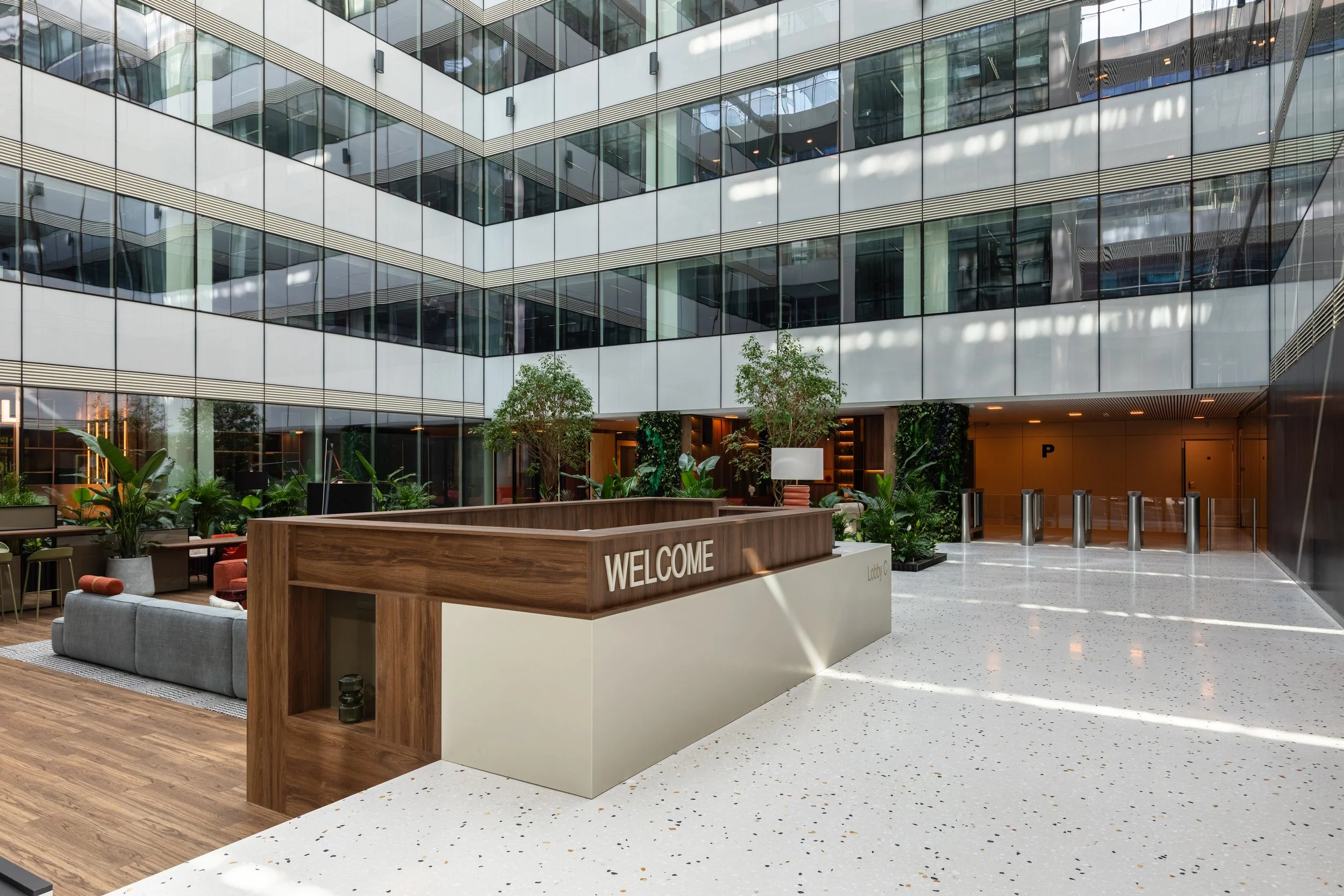 Modern office lobby with waiting area, reception desk labeled "Welcome," indoor plants, large glass windows reflecting the exterior, and security gates at the entrance.