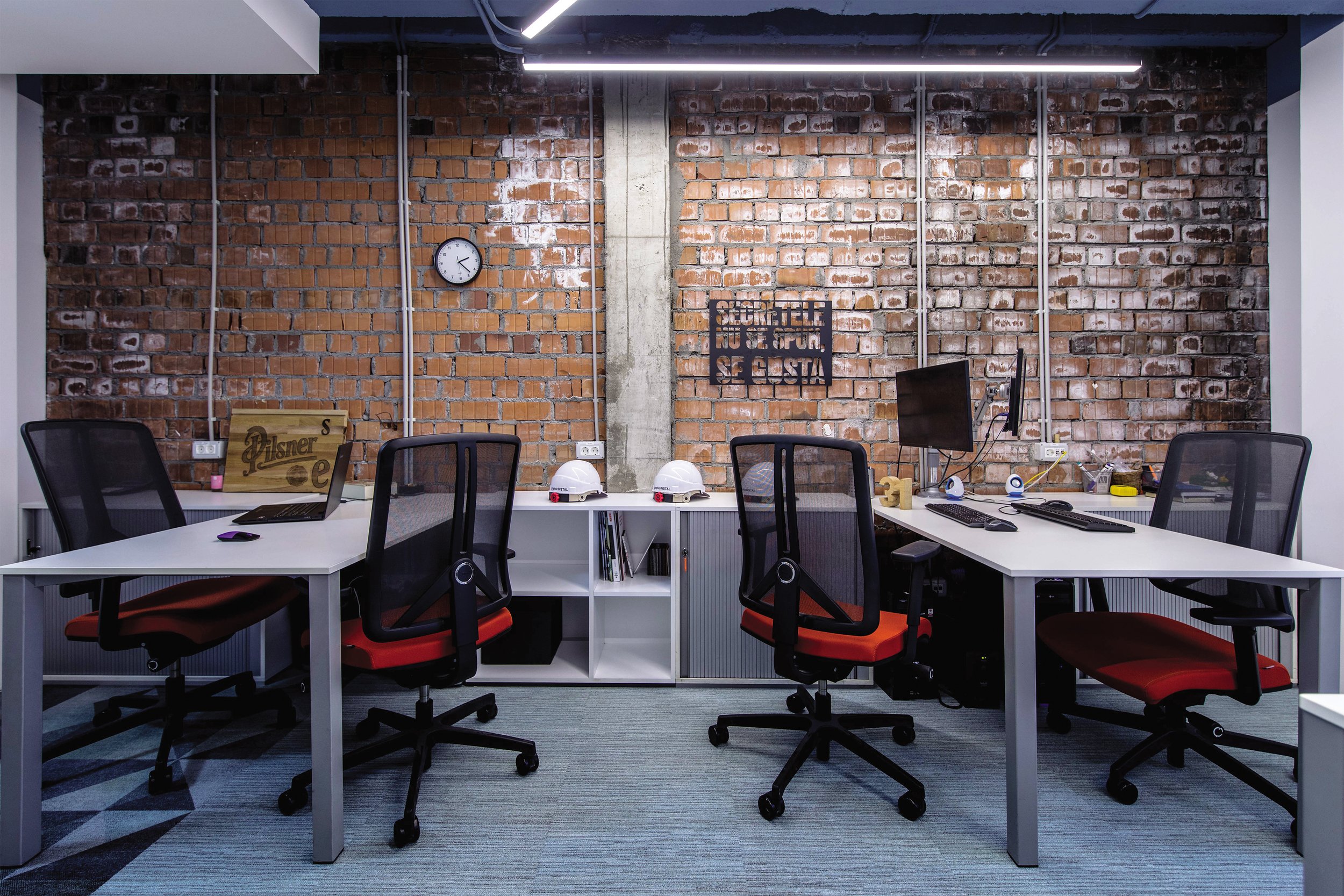 Modern office workspace with four black mesh office chairs with red cushions around two white desks, a brick wall background, white storage units underneath the desks, with two white hard helmets, books, computers, and office supplies. A clock on the wall shows 1:10, and there is a sign that says in Spanish, "Secreto no es su hermano, se gosta."