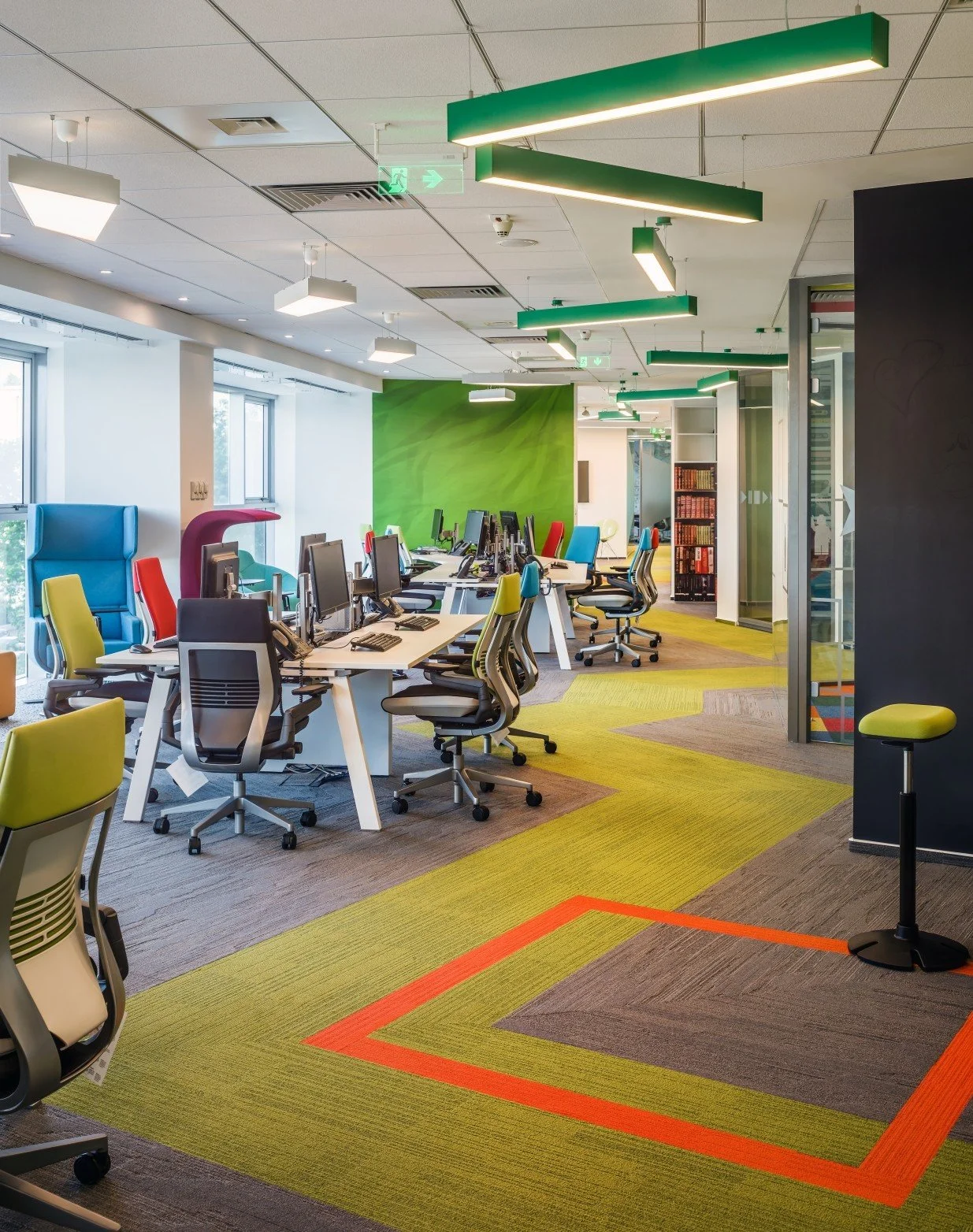 Modern office with colorful chairs, desktop computers, and green wall in an open-plan workspace.
