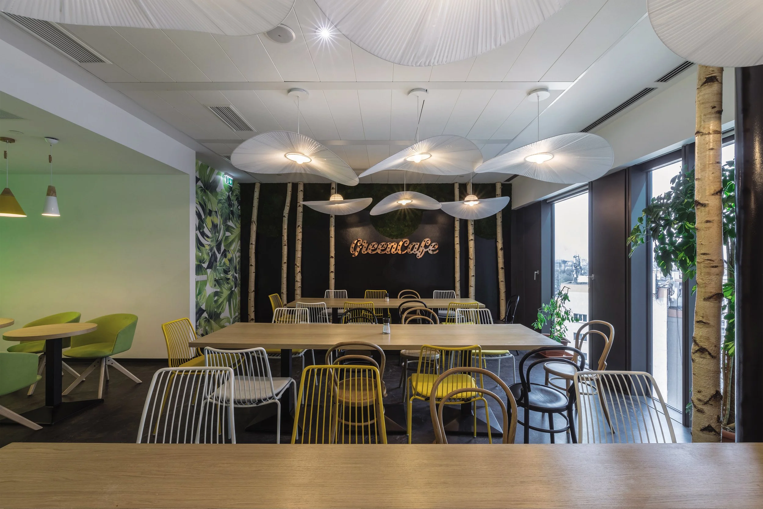 Interior of a Modern cafe with wooden tables, colorful chairs, decorative hanging lights, and natural light from large windows; sign on the wall reads 'Green Cafe'.