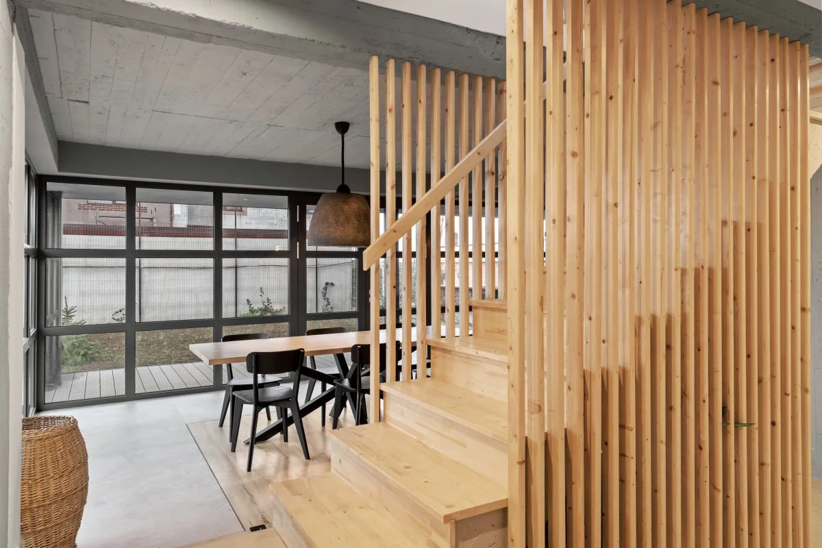 Interior of a modern home with a wooden staircase, wooden slat wall, dining area with black chairs around a light wood table, large glass sliding door, concrete ceiling, and a hanging pendant light.