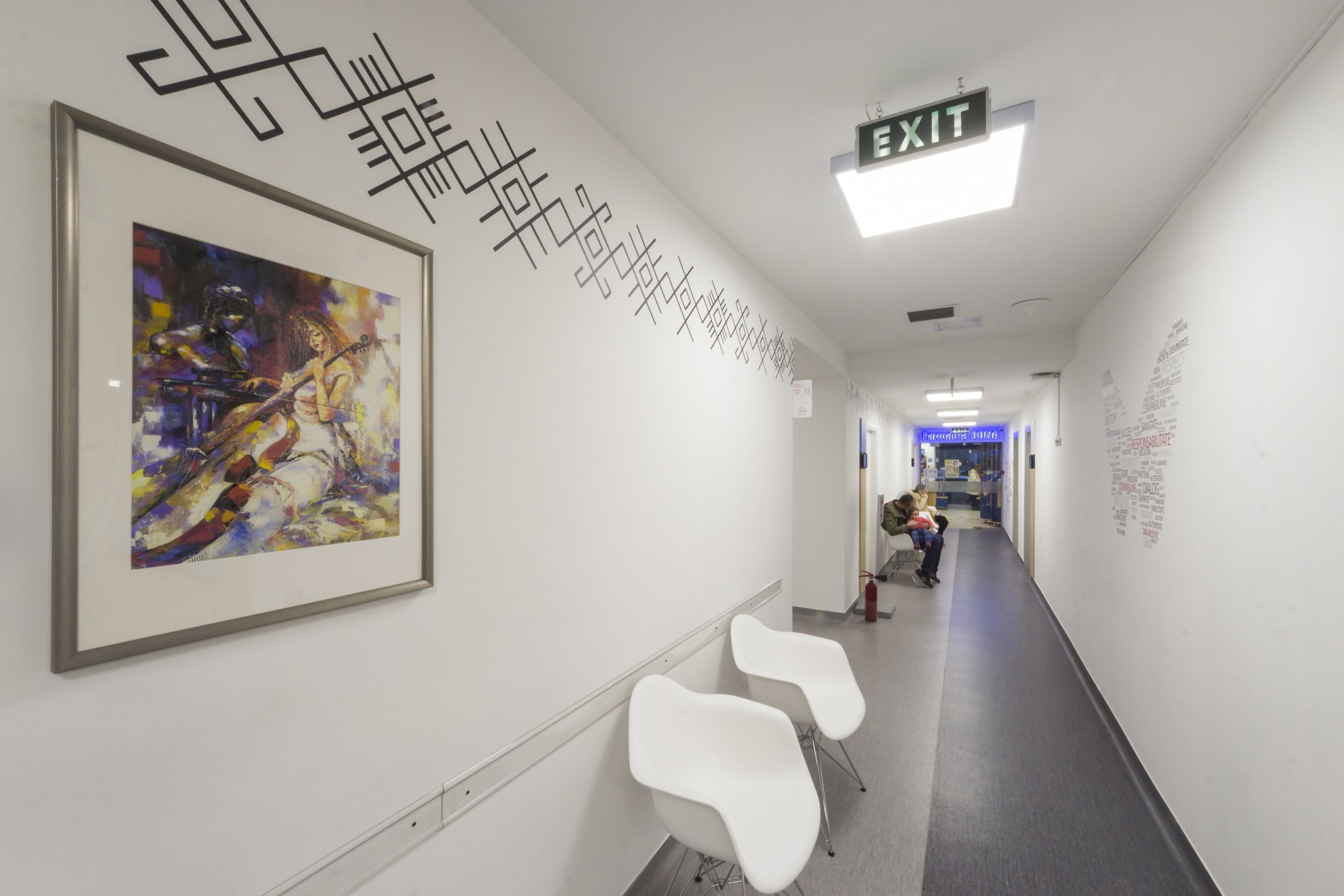 Hospital corridor with white walls, colorful abstract painting on the left wall, white chairs on the right side, an exit sign on the ceiling, and a person sitting with a child.