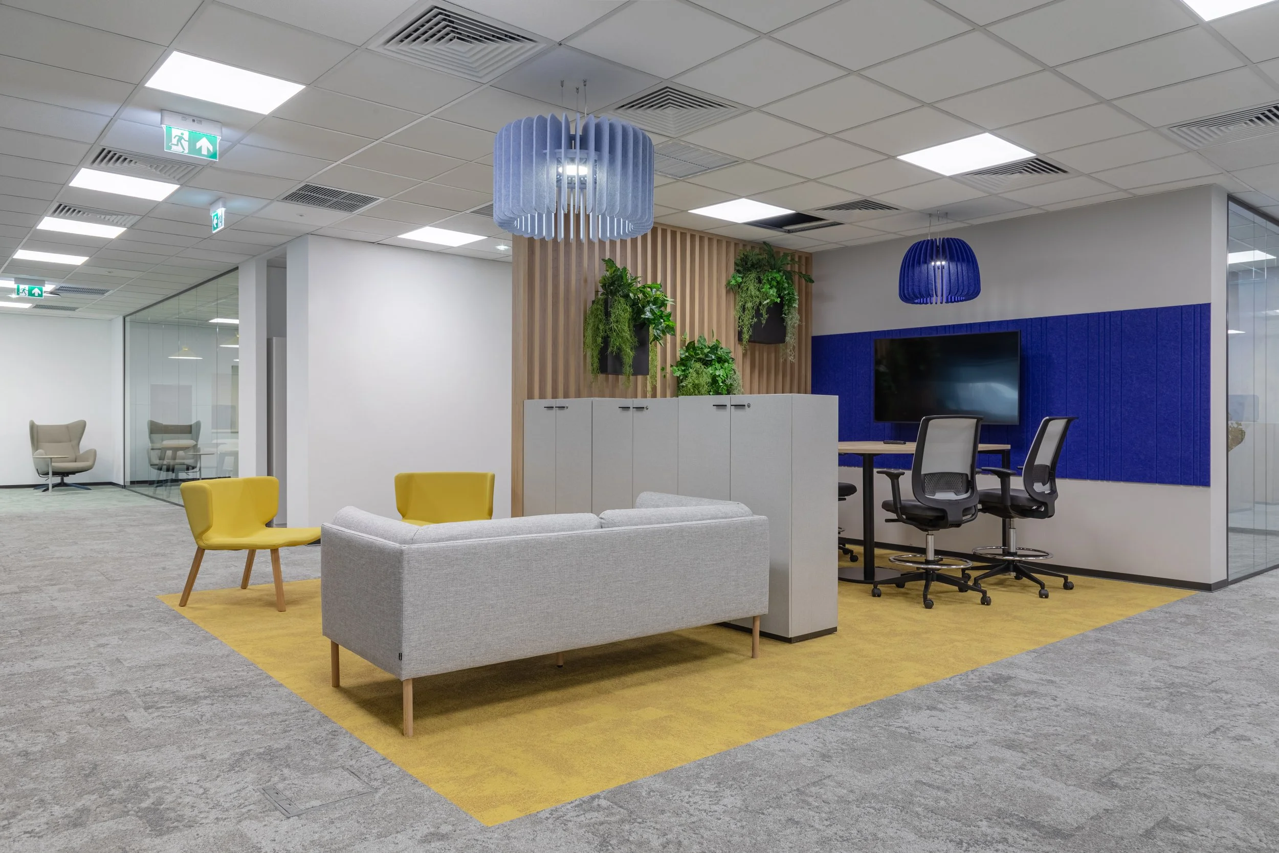 Modern office lounge with yellow chairs, a gray sofa, and a meeting area with chairs around a table. Decorative plants are hung on a wooden slat wall. Two hanging blue pendant lights and a blue acoustic panel behind a large monitor are visible.