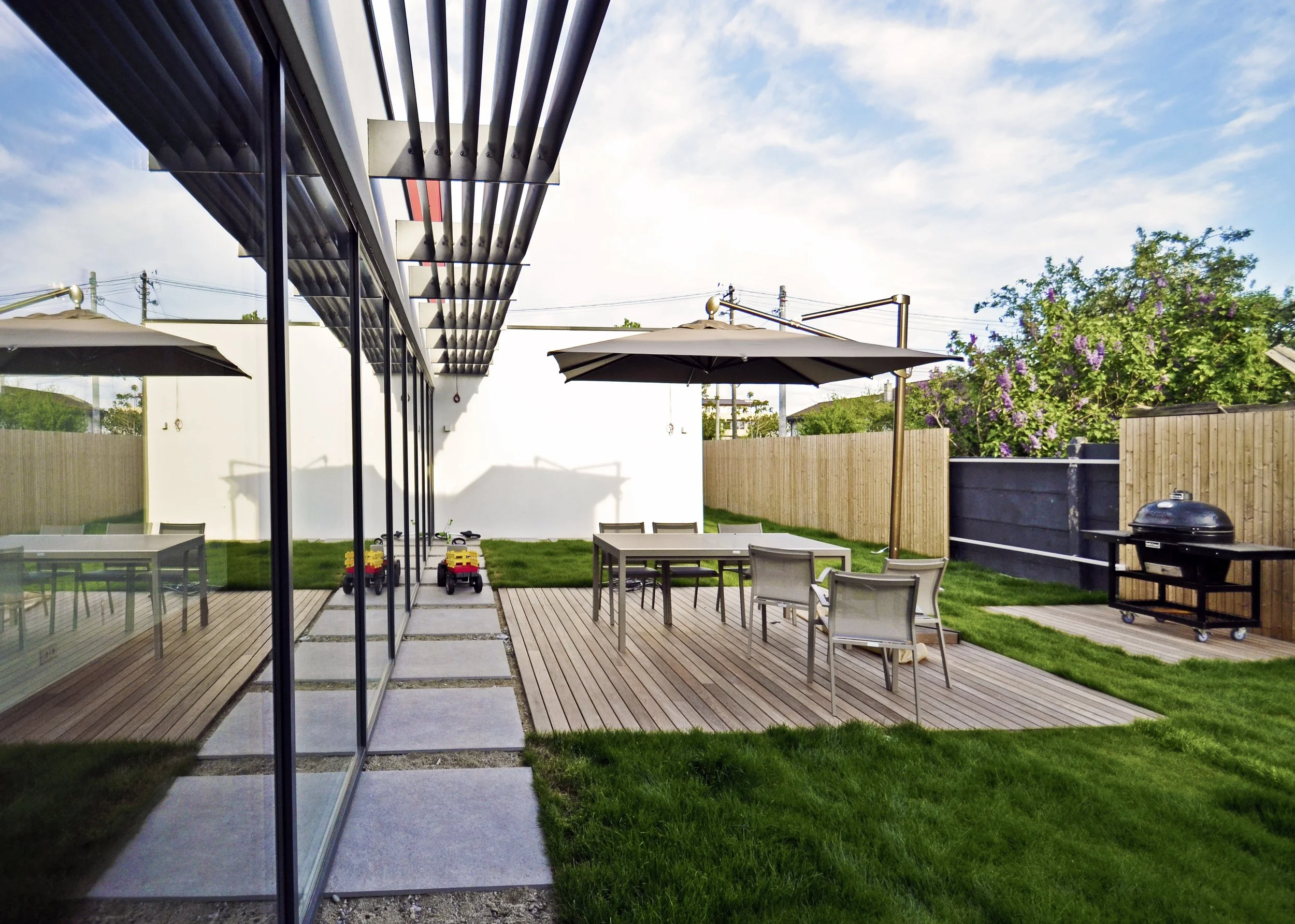 A backyard patio with outdoor dining table and chairs, large umbrellas, a barbecue grill, and a lawn area with a wooden privacy fence, flowering trees, and a cloudy sky.