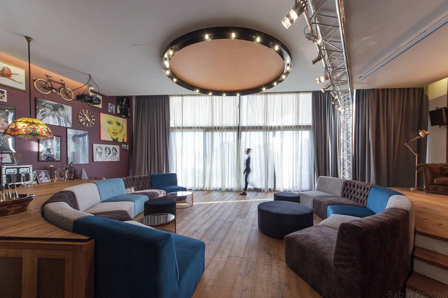 A spacious lounge with large windows, a circular ceiling light, velvet couches, and wall art including a vintage bicycle and portraits, with a person walking in front of the windows.