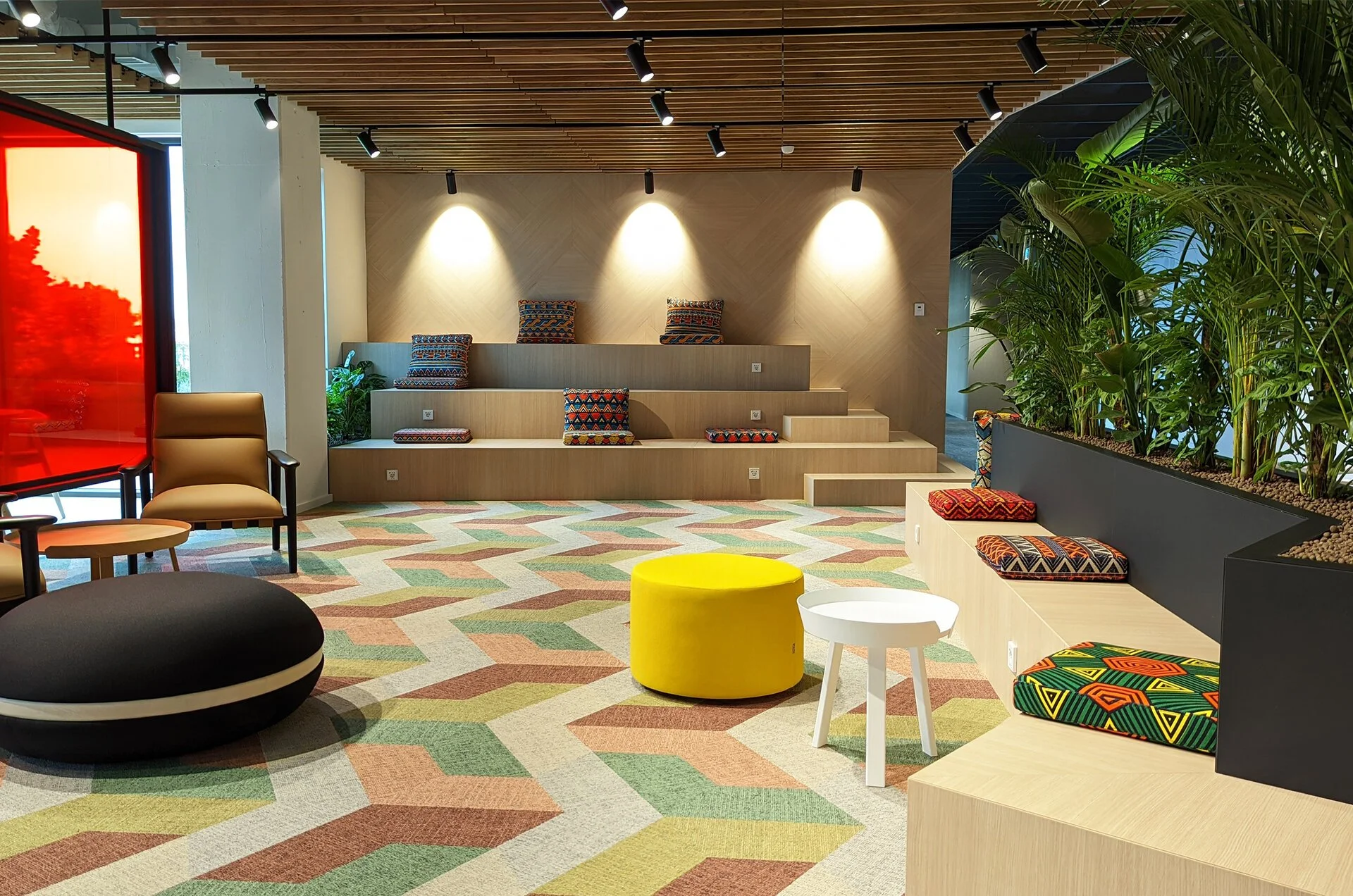 Modern indoor lounge area with tiered seating, colorful patterned cushions, two bags, and vibrant geometric patterned carpet, illuminated by ceiling spotlights, with a large planter and plants on the right.