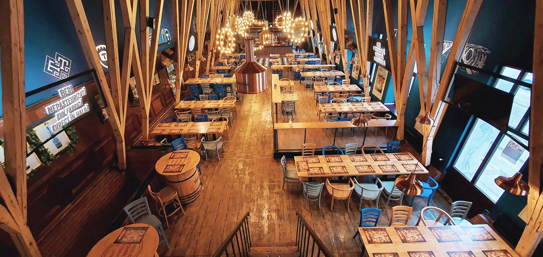 Empty restaurant with wooden tables and chairs, large copper light fixtures, and large windows; the interior has a rustic, modern aesthetic.