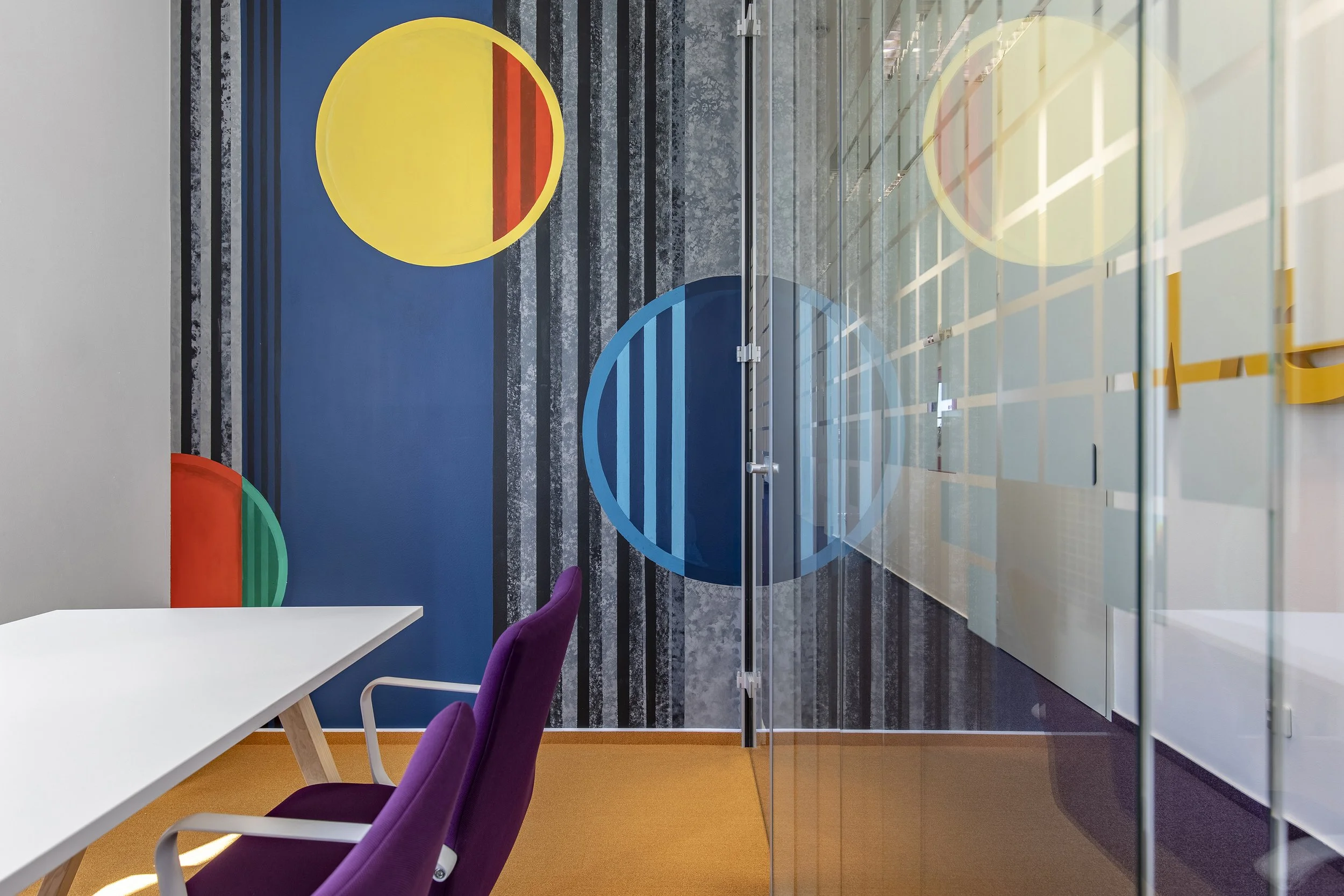 Modern office conference room with a white table, purple chairs, and a glass wall with a colorful abstract mural of circles and vertical stripes.
