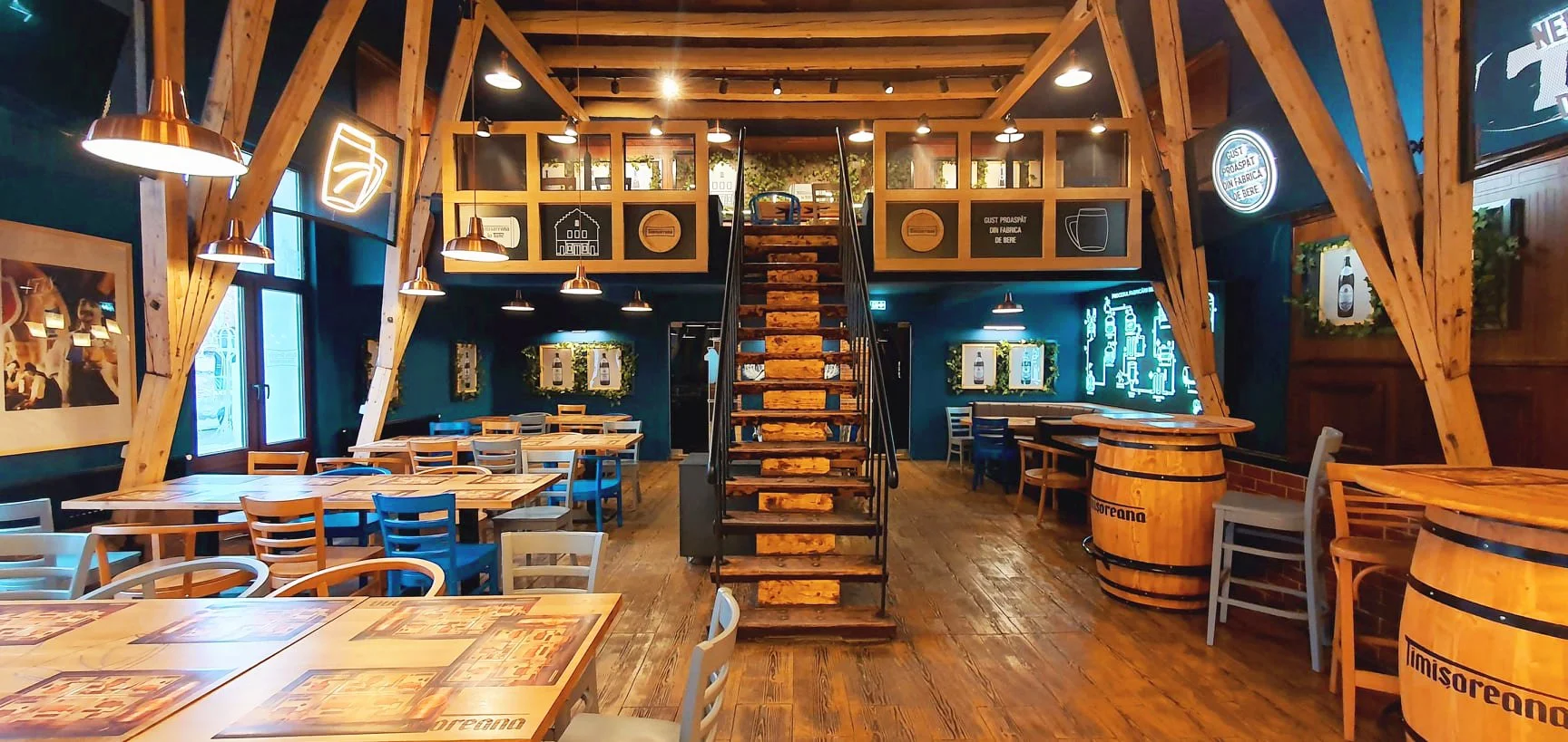 Inside a rustic restaurant or brewery with wooden beams, stairs, and barrels, modern lighting, framed photos, and neon signs on dark green and wood-paneled walls.