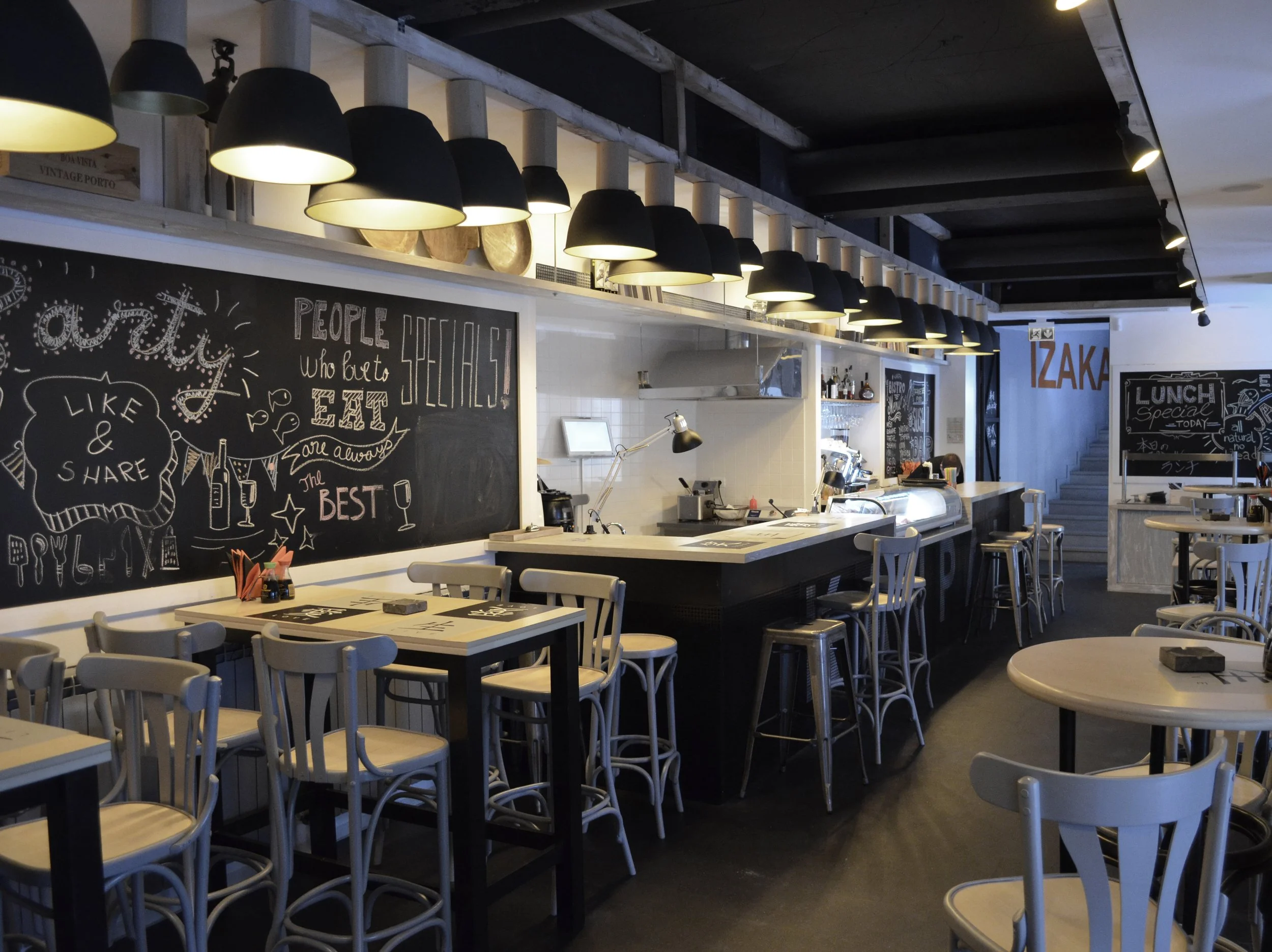 Interior of a modern restaurant with white chairs, black tables, and a black chalkboard wall with handwritten menus and decorative drawings.