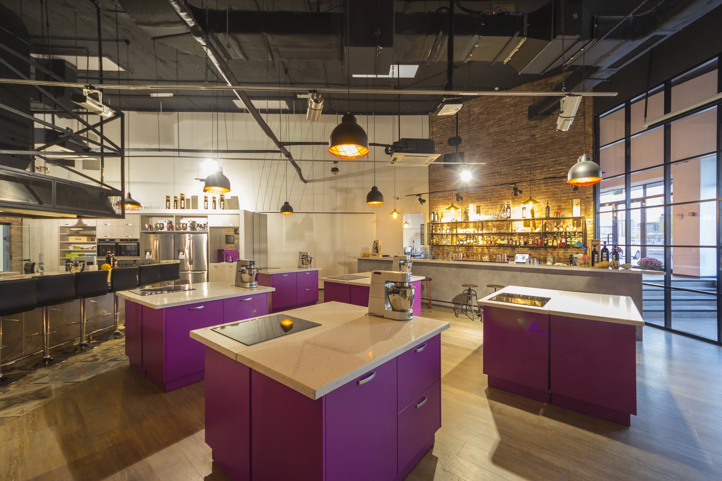 Interior of a modern, well-lit commercial kitchen or bar area with purple kitchen islands, black pendant lights, a bar with bottles and bar stools, and large windows letting in natural light.