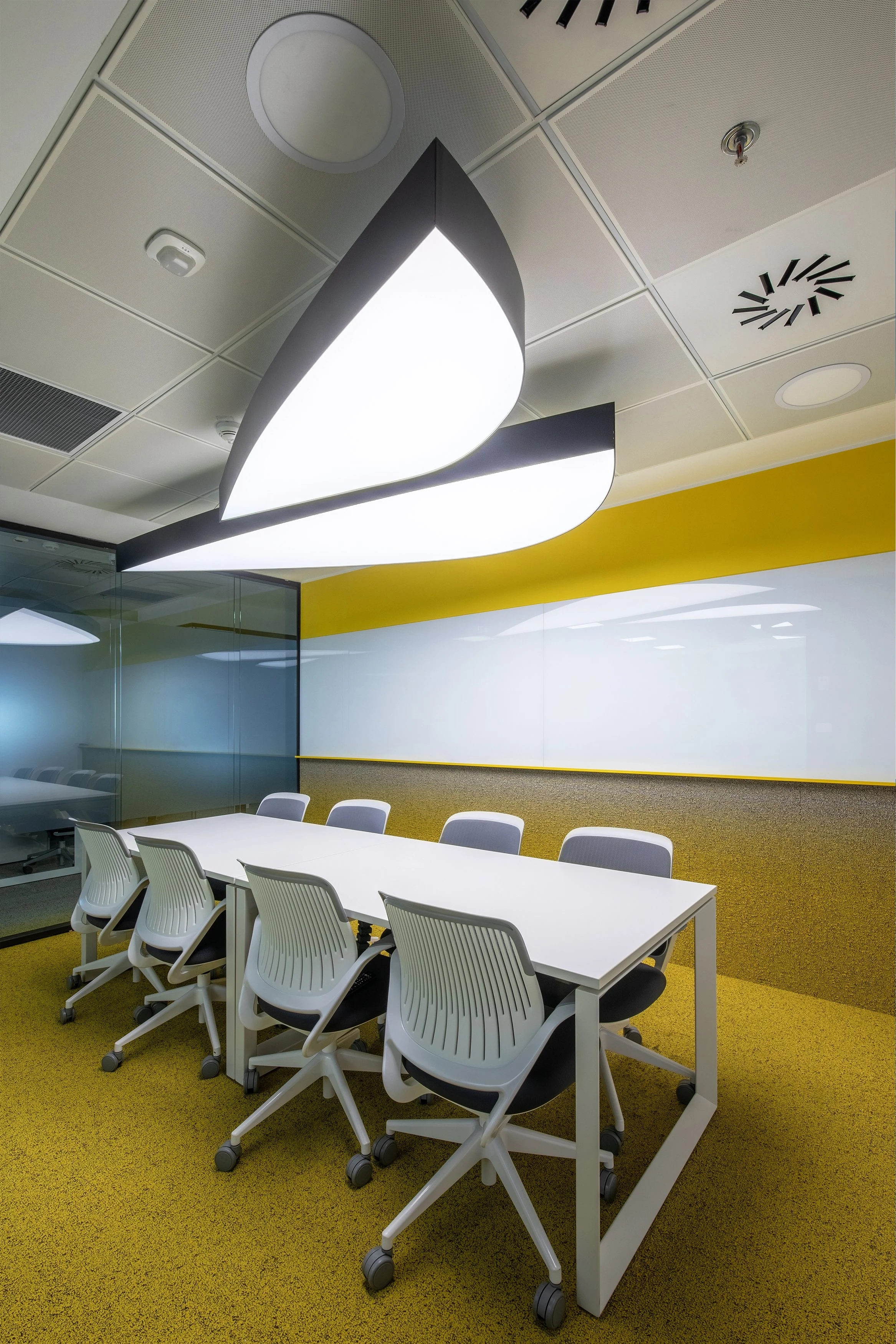 A modern conference room with a long white table surrounded by eight white ergonomic office chairs with black seats and gray backs. Above, a large leaf-shaped light fixture is mounted on the ceiling, which features a drop ceiling with acoustic tiles, a smoke detector, a clock, and a fire sprinkler. The room has a yellow accent wall behind the table, with a frosted glass wall to the left.