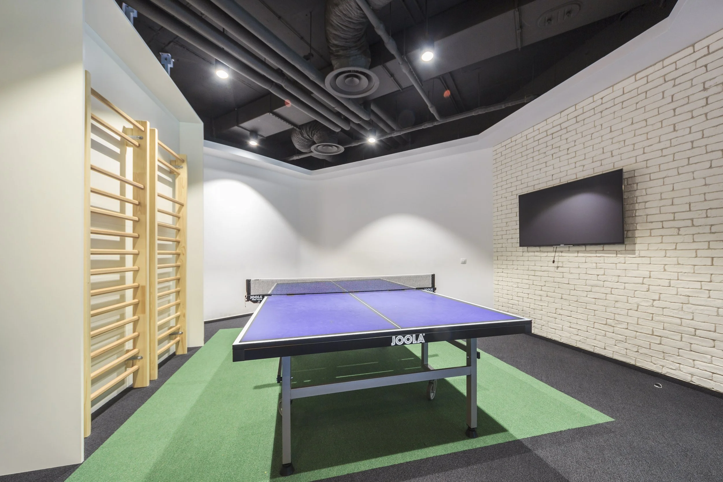 Indoor recreational space featuring a blue ping pong table, green and black carpeted flooring, a flat-screen TV mounted on a white brick wall, and wooden wall bars, with black ceiling and ceiling lights.
