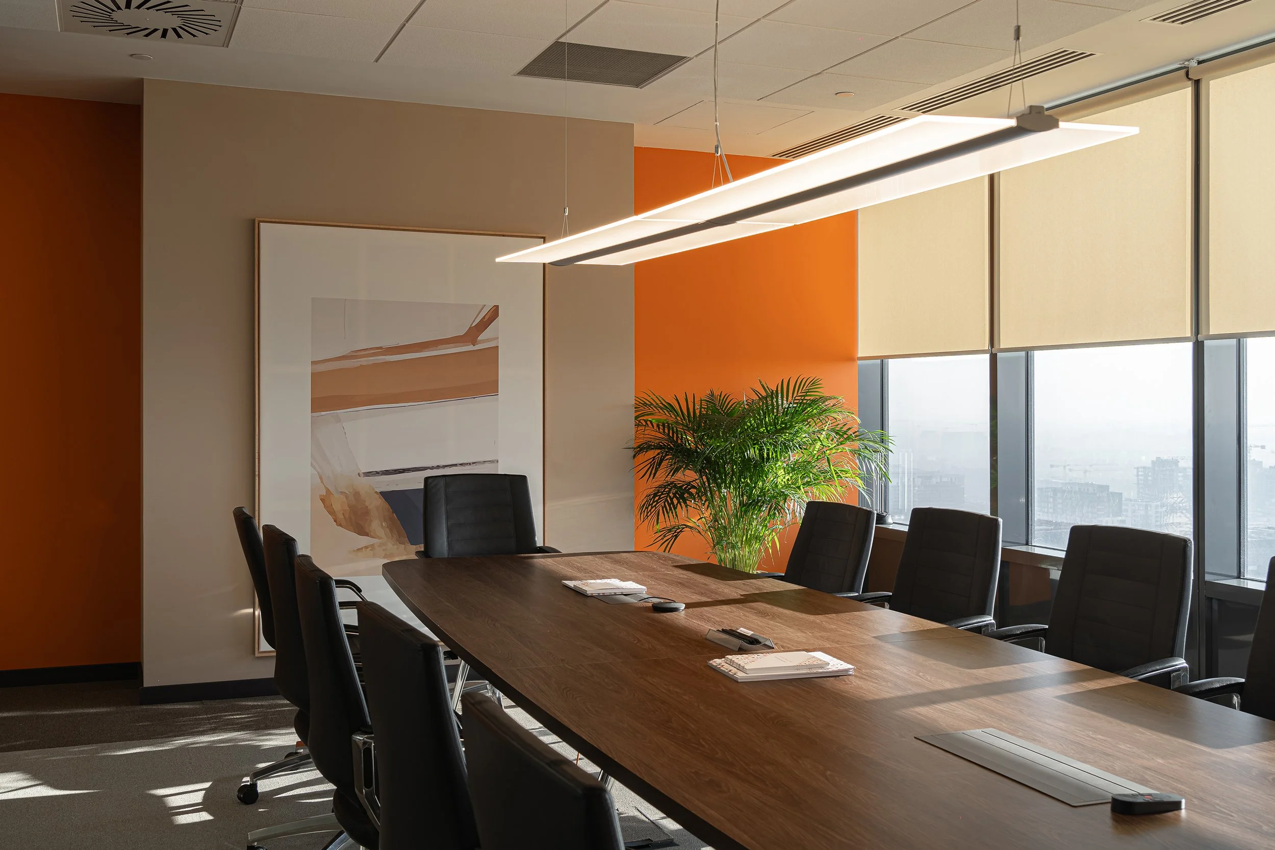 Modern conference room with a long wooden table, black leather chairs, large windows with blinds, a large plant, and abstract artwork on the wall.
