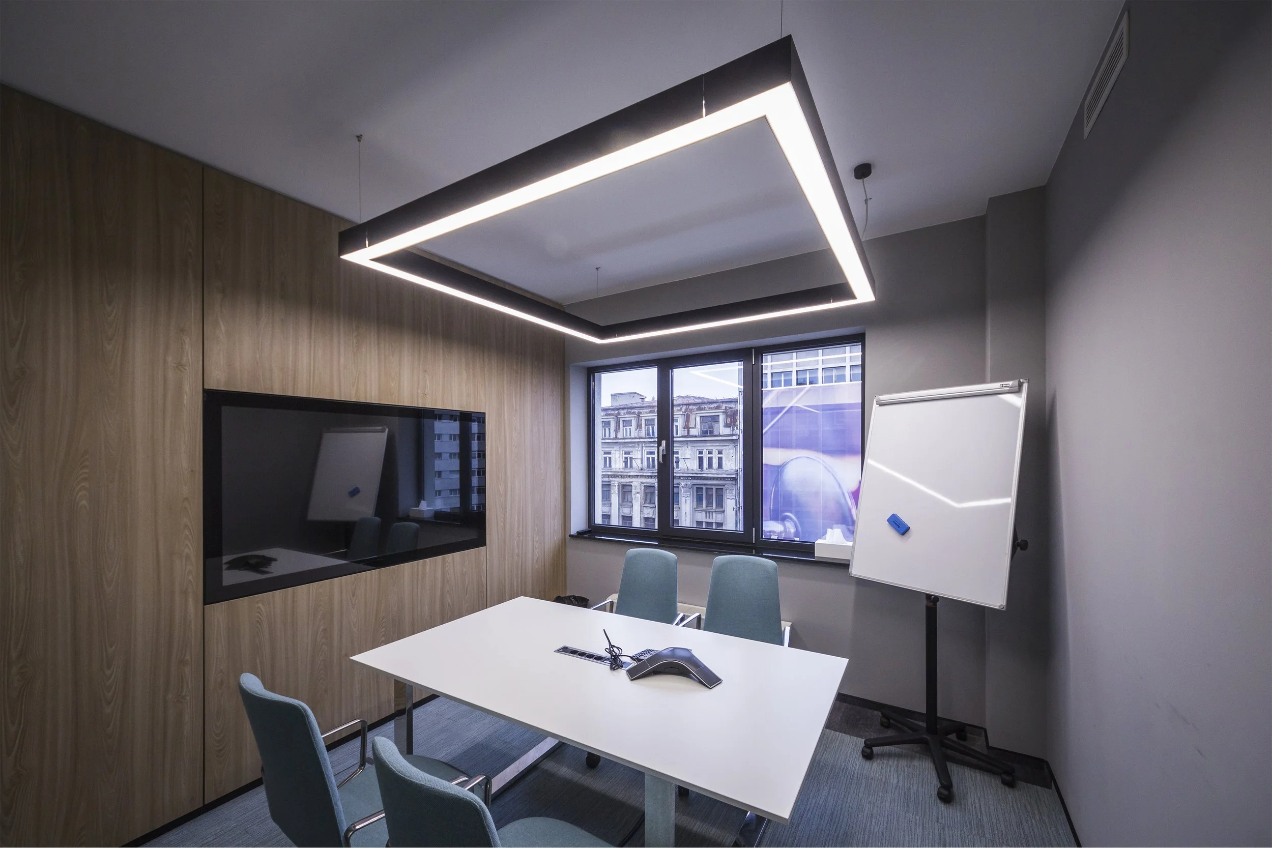 Modern office room with a white table, four chairs, a whiteboard on a stand, a large window showing city buildings, a wooden accent wall with a flat screen, and a rectangular ceiling light