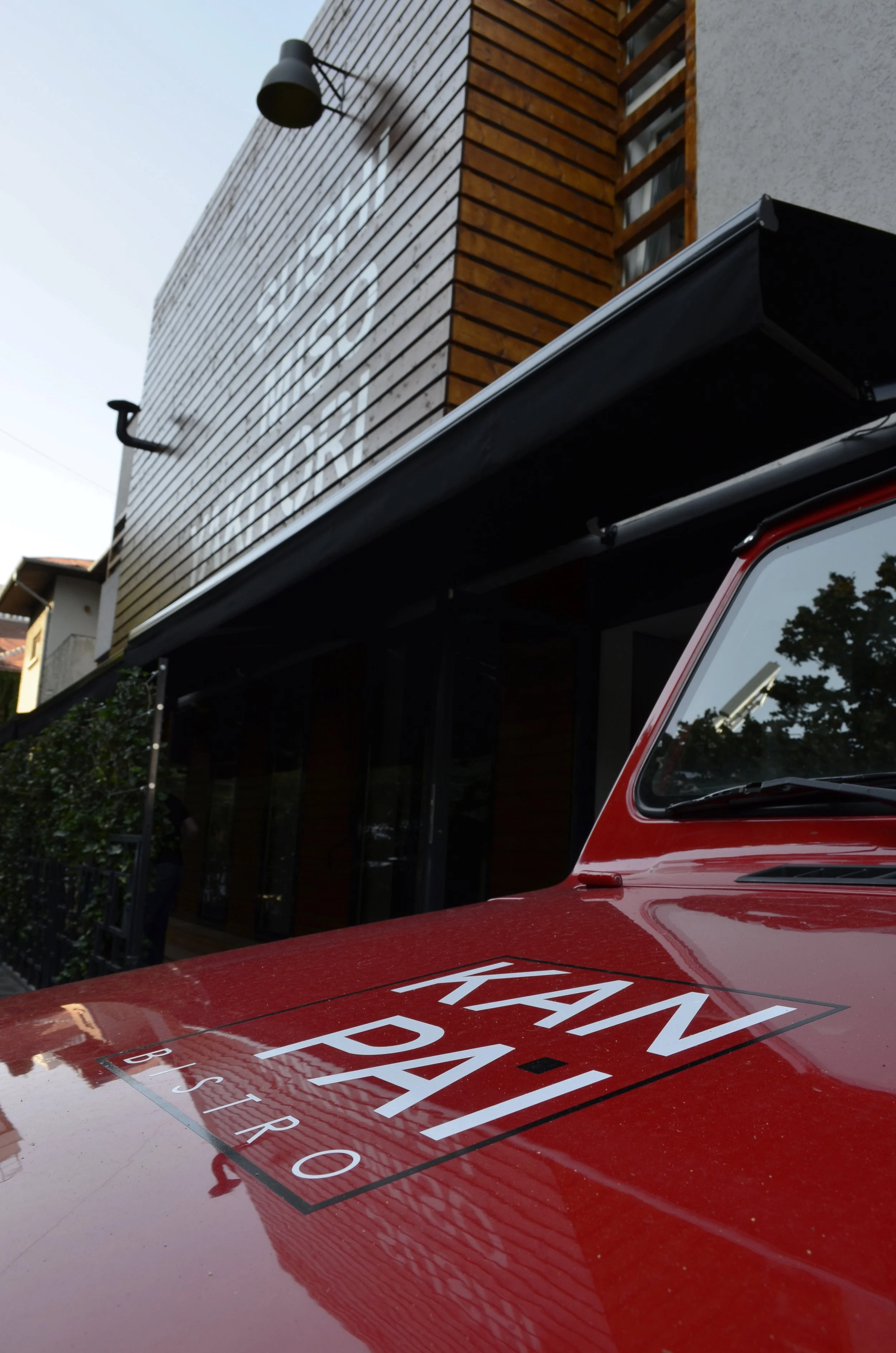 Close-up view of a red vehicle with a sign on its hood that reads 'KANADA BISTRO,' parked in front of a modern building with wooden siding and a black awning.