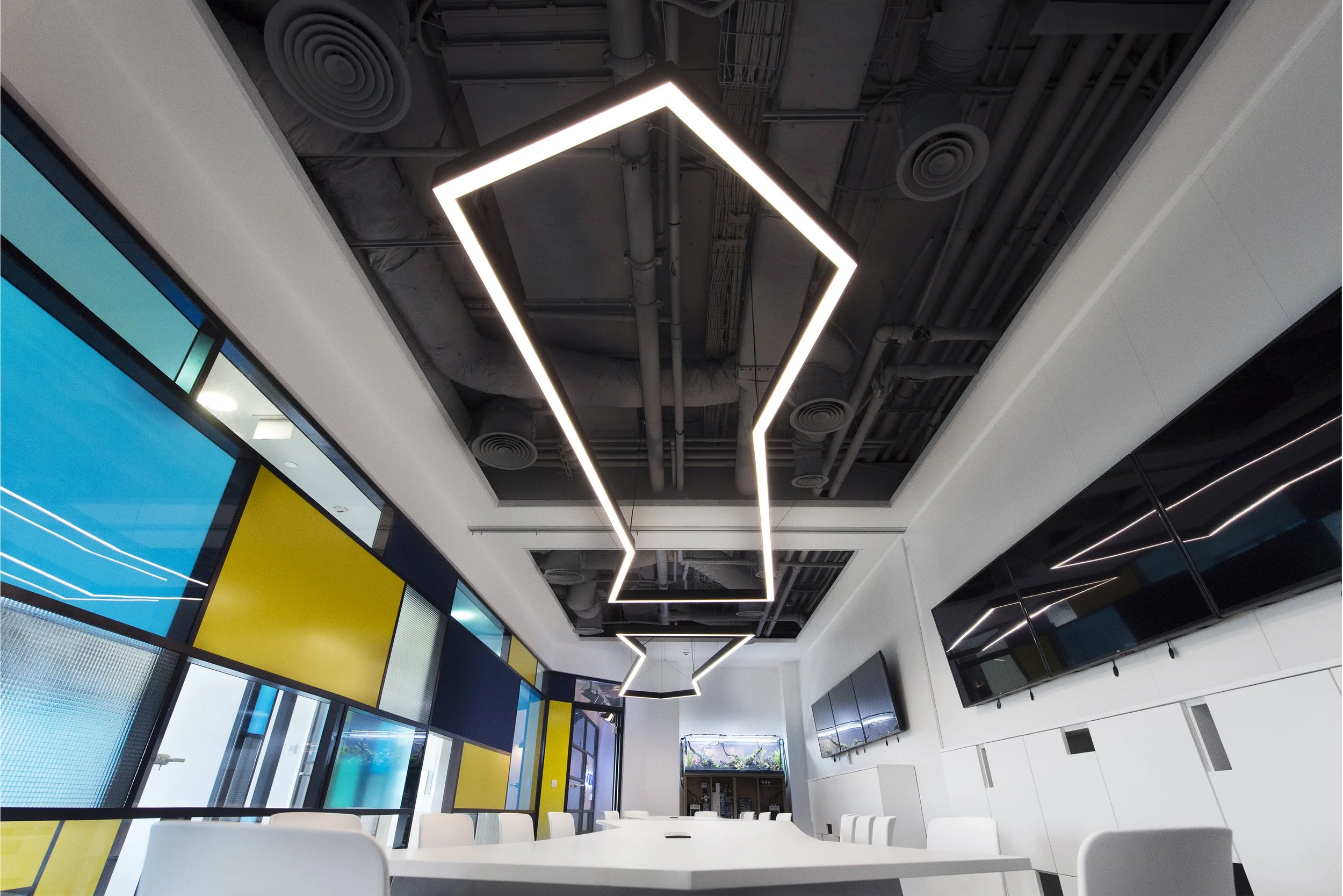 Modern conference room with white table, white chairs, wall-mounted TV screens, colorful window panels, and geometric ceiling lighting fixture.