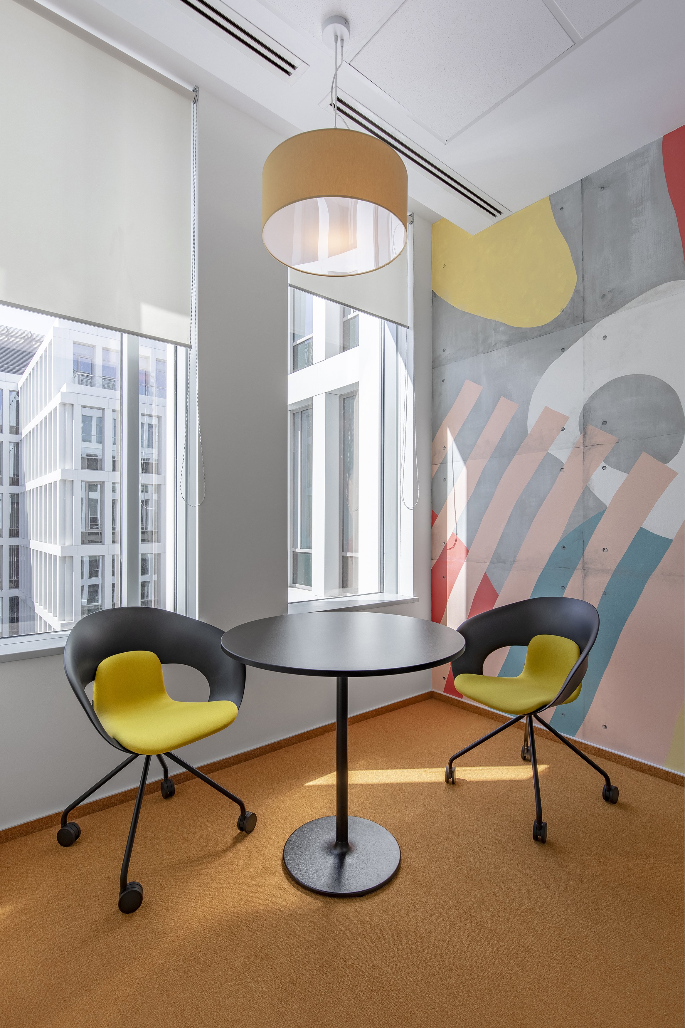 Small meeting room with two black chairs with yellow cushions, black round table, colorful abstract wall mural, large windows with blinds, orange carpet, and beige pendant light.