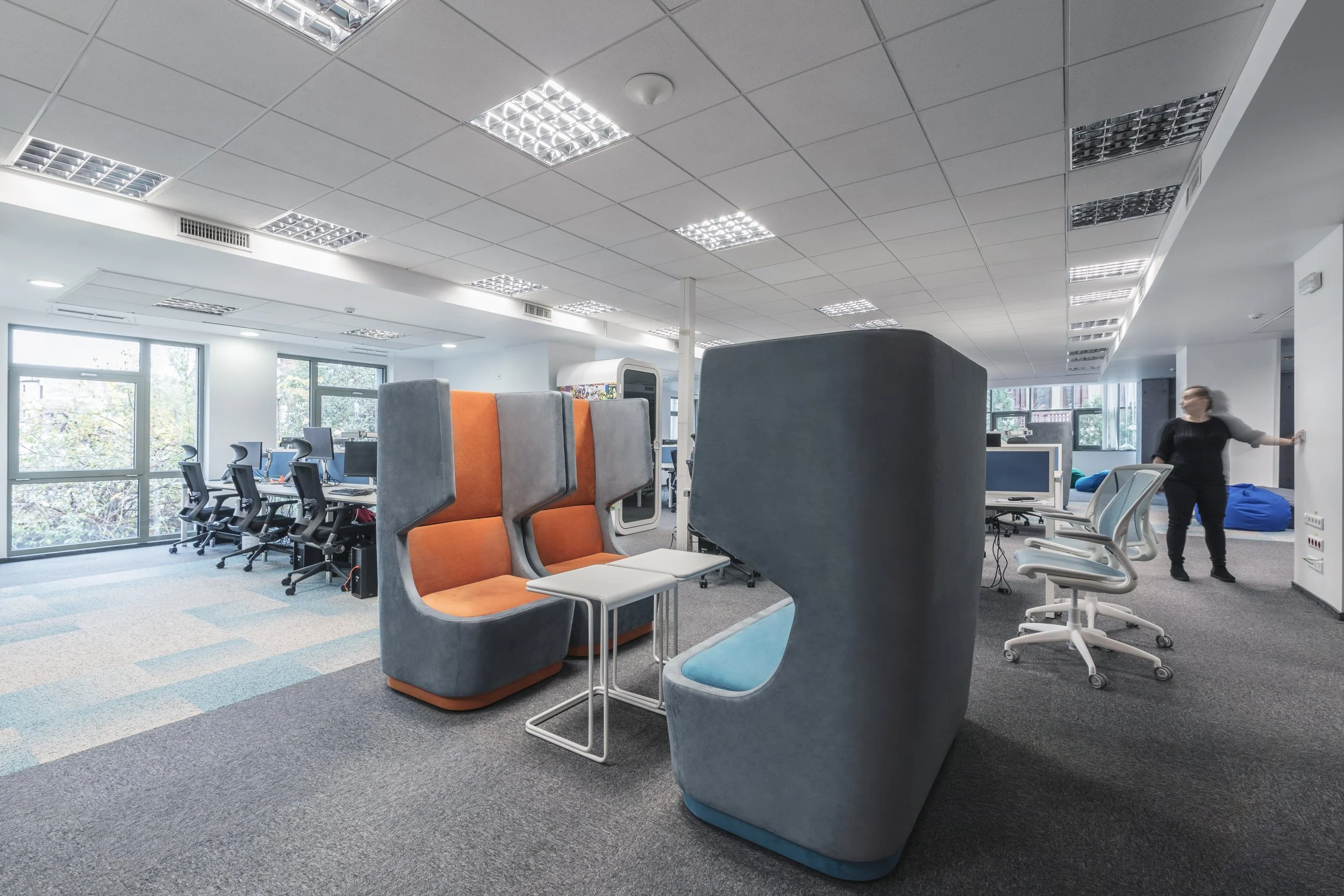 Open office space with workstations, chairs, and colorful privacy pods, illuminated by ceiling lights and natural light from large windows.