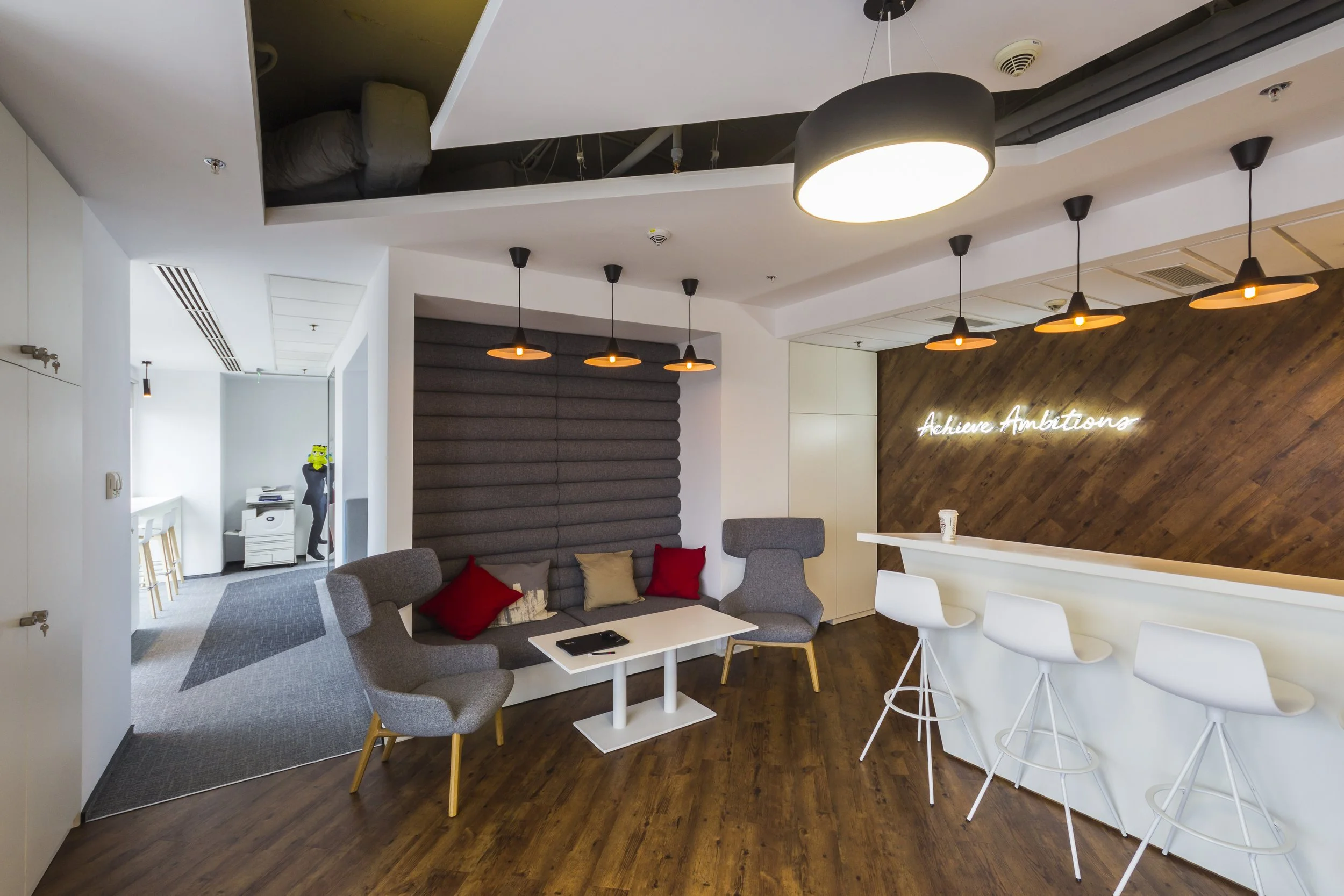 Modern coffee shop interior with gray lounge chairs, a white bar counter with three white bar stools, decorative pillows on the chairs, a dark wood accent wall with a neon sign reading 'Achieve Ambitions', pendant lighting, and a small seating area with a white table.