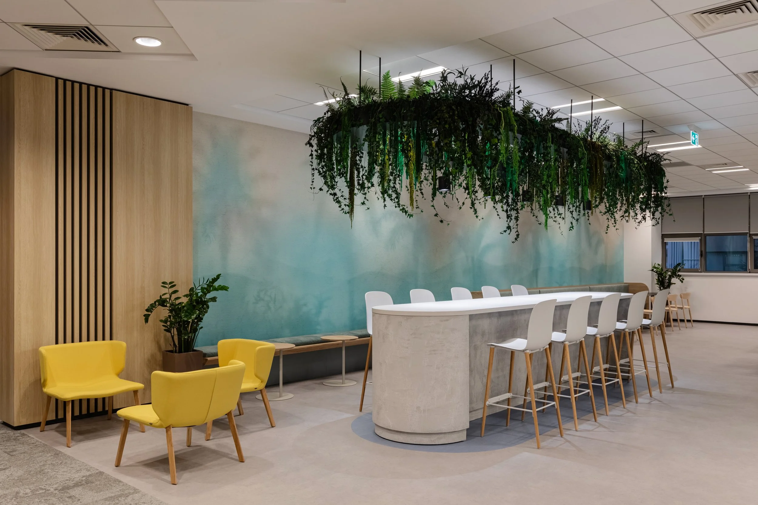 Modern office lounge area featuring yellow chairs, white bar stools at a curved concrete counter, green plants, and a hanging garden with lush greenery overhead.