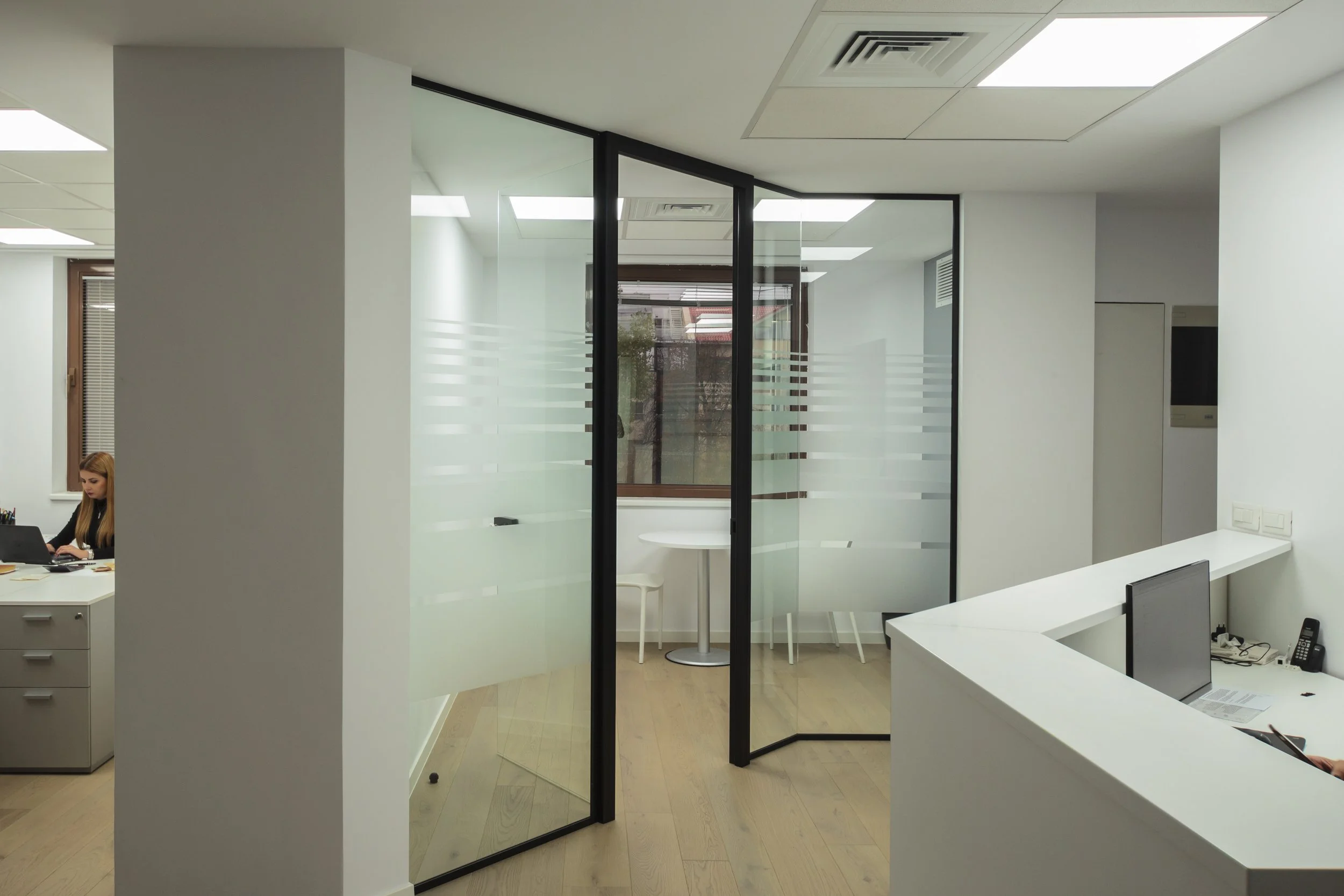 Modern office space with glass partitions, a reception desk with computer, and an enclosed seating area with a small table and chairs