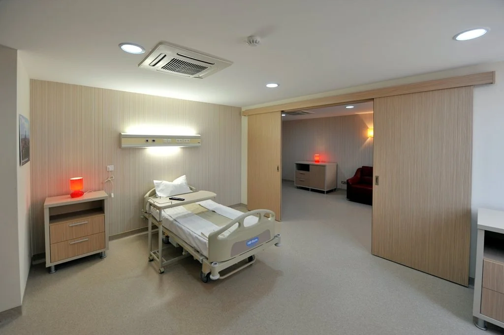 Hospital room with a bed, nightstand, and sliding door leading to a sitting area with a red chair and nightlight.