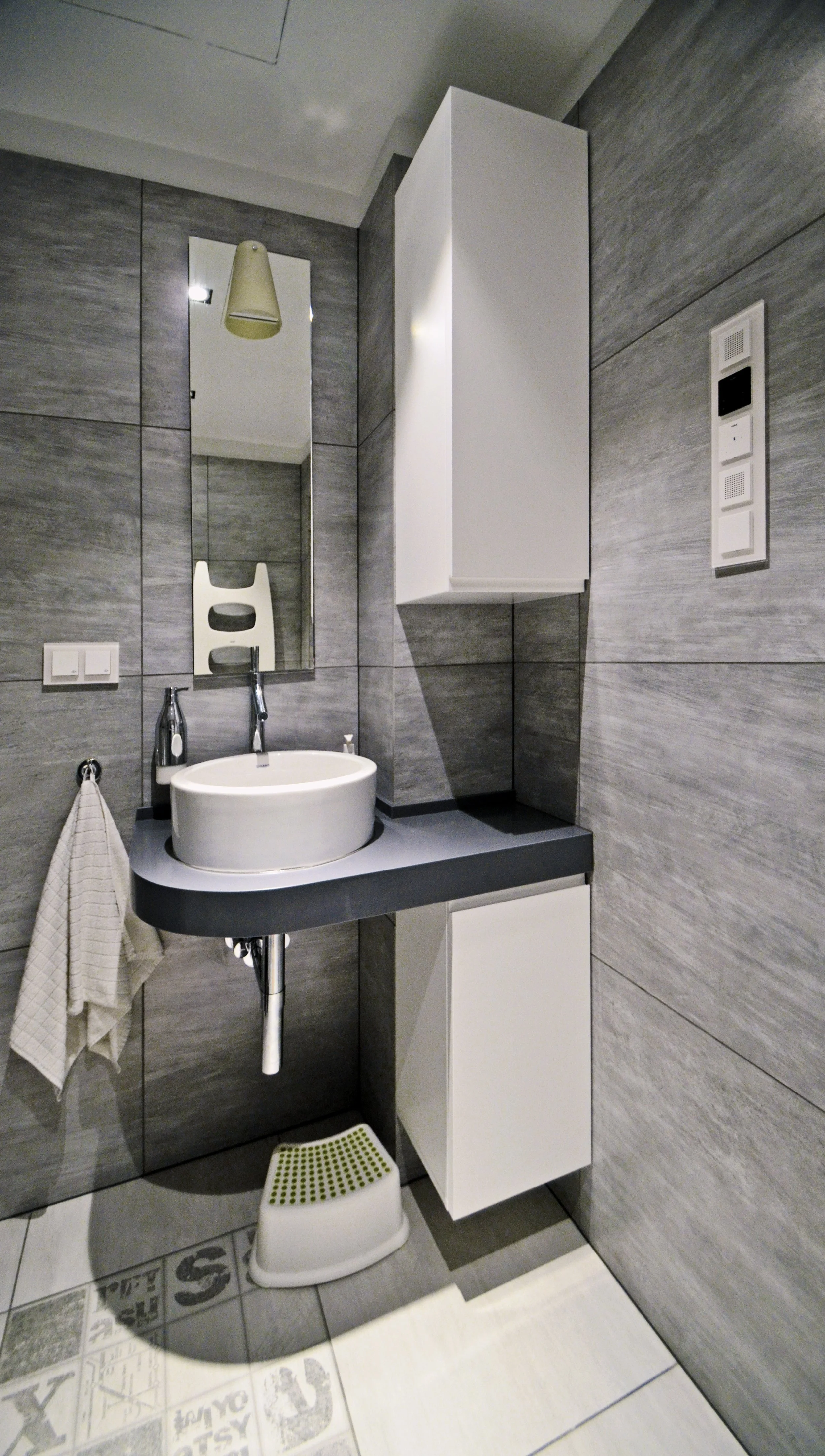 Modern bathroom with gray tiled walls, a round white vessel sink on a dark countertop, a mirror, a towel hanging, a white cabinet, and a small step stool with green circles.