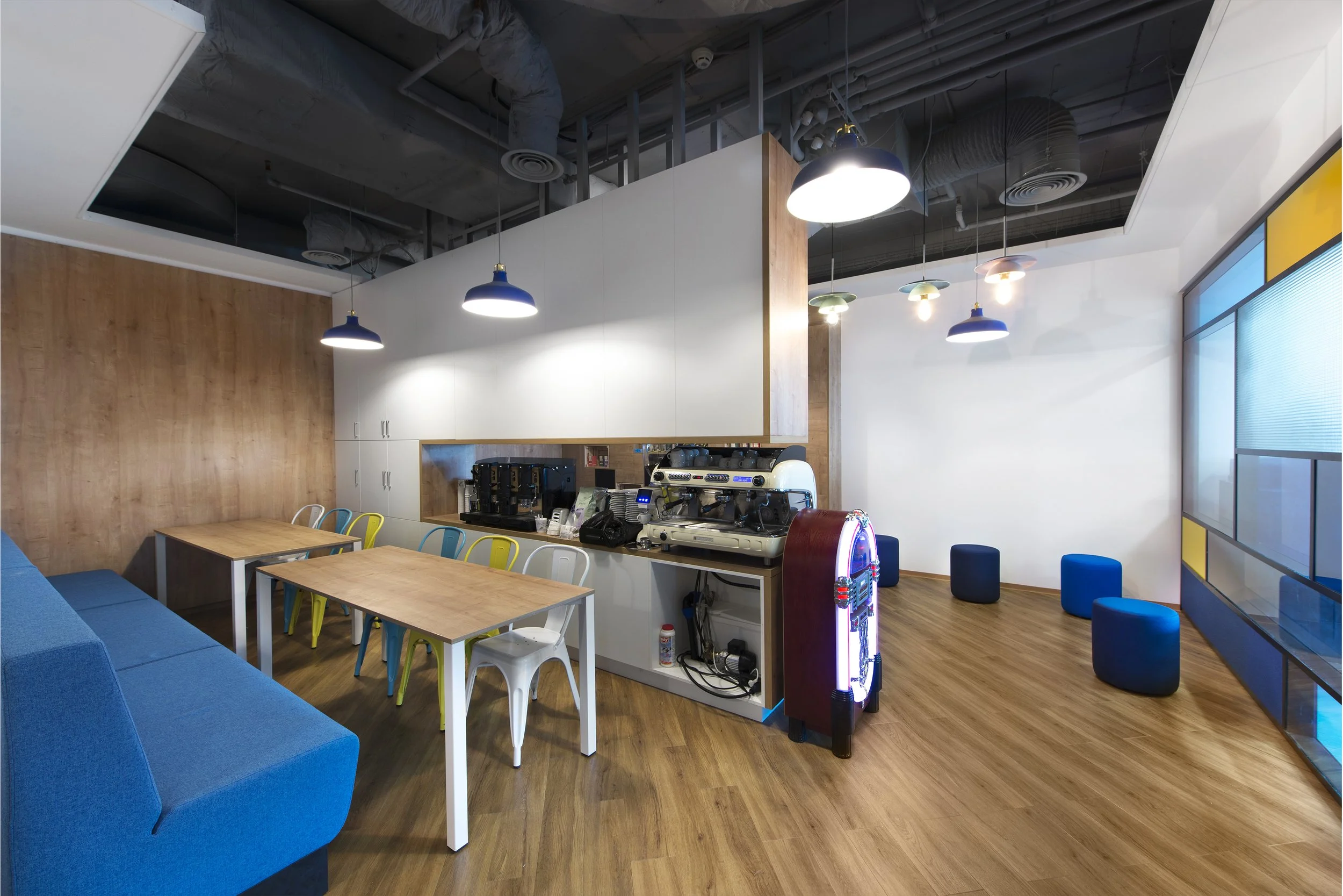 Modern coffee shop with wooden tables, colorful chairs, blue seating, and coffee brewing equipment on a counter, illuminated by hanging pendant lights.