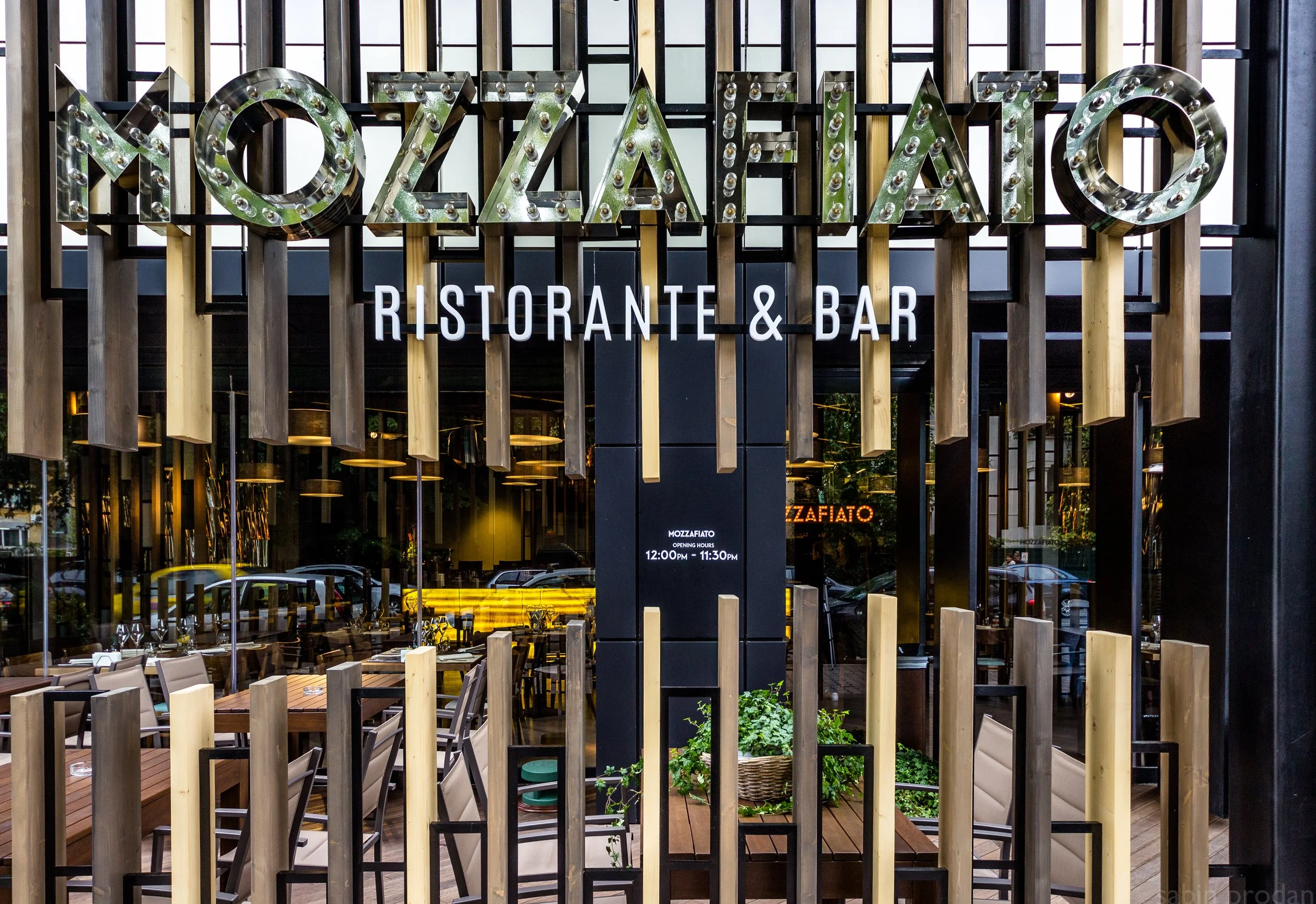Exterior view of Mozzafiato Ristorante & Bar with modern design, featuring wood and black metal elements, large glass windows, outdoor seating, and a decorative sign with lights.