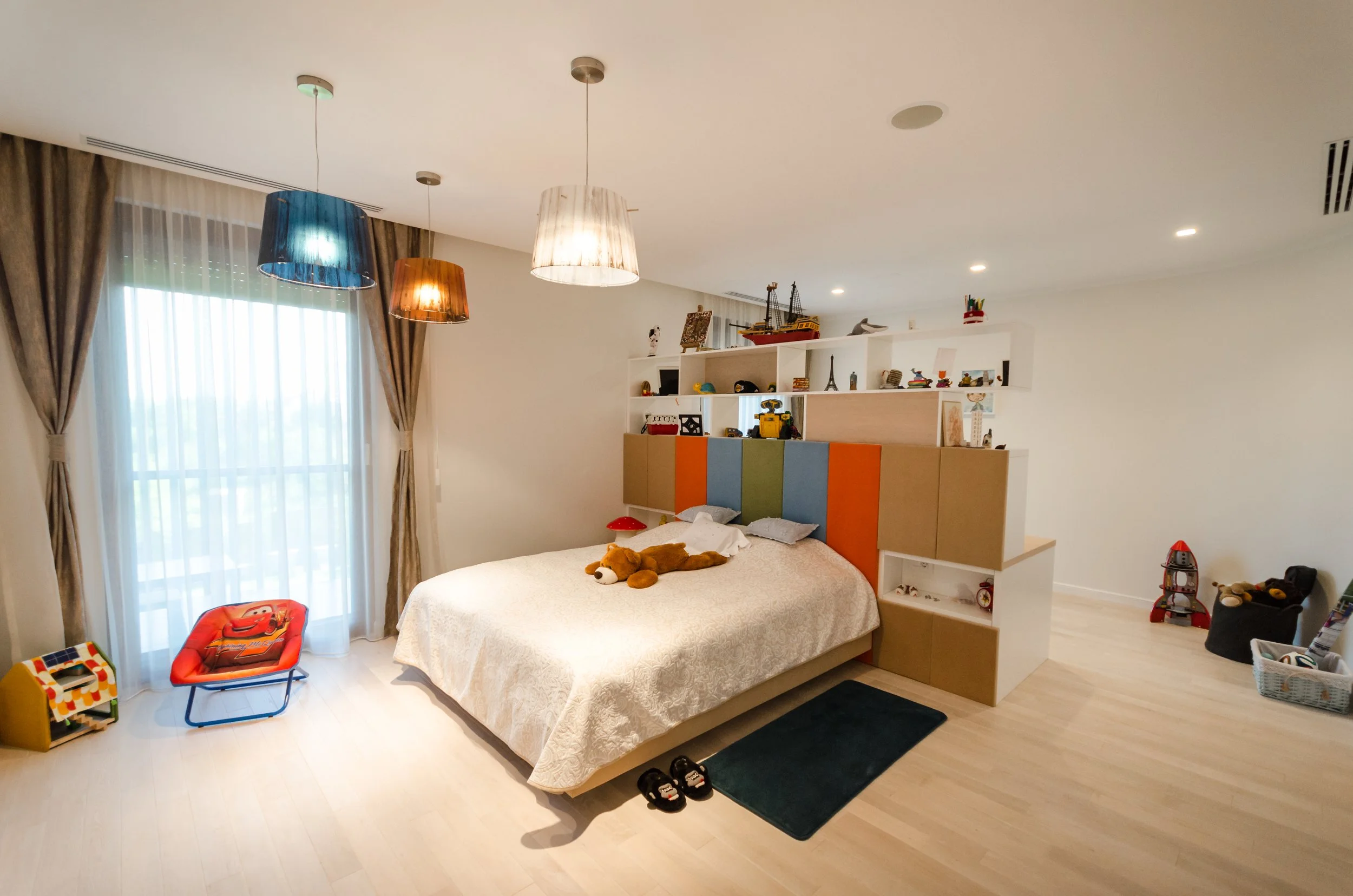 A bedroom with a bed, colorful headboard, and stuffed toys. There are large window curtains with sheer curtains, hanging pendant lights, and a toy chair. Shelves above the bed hold toys and decorations.