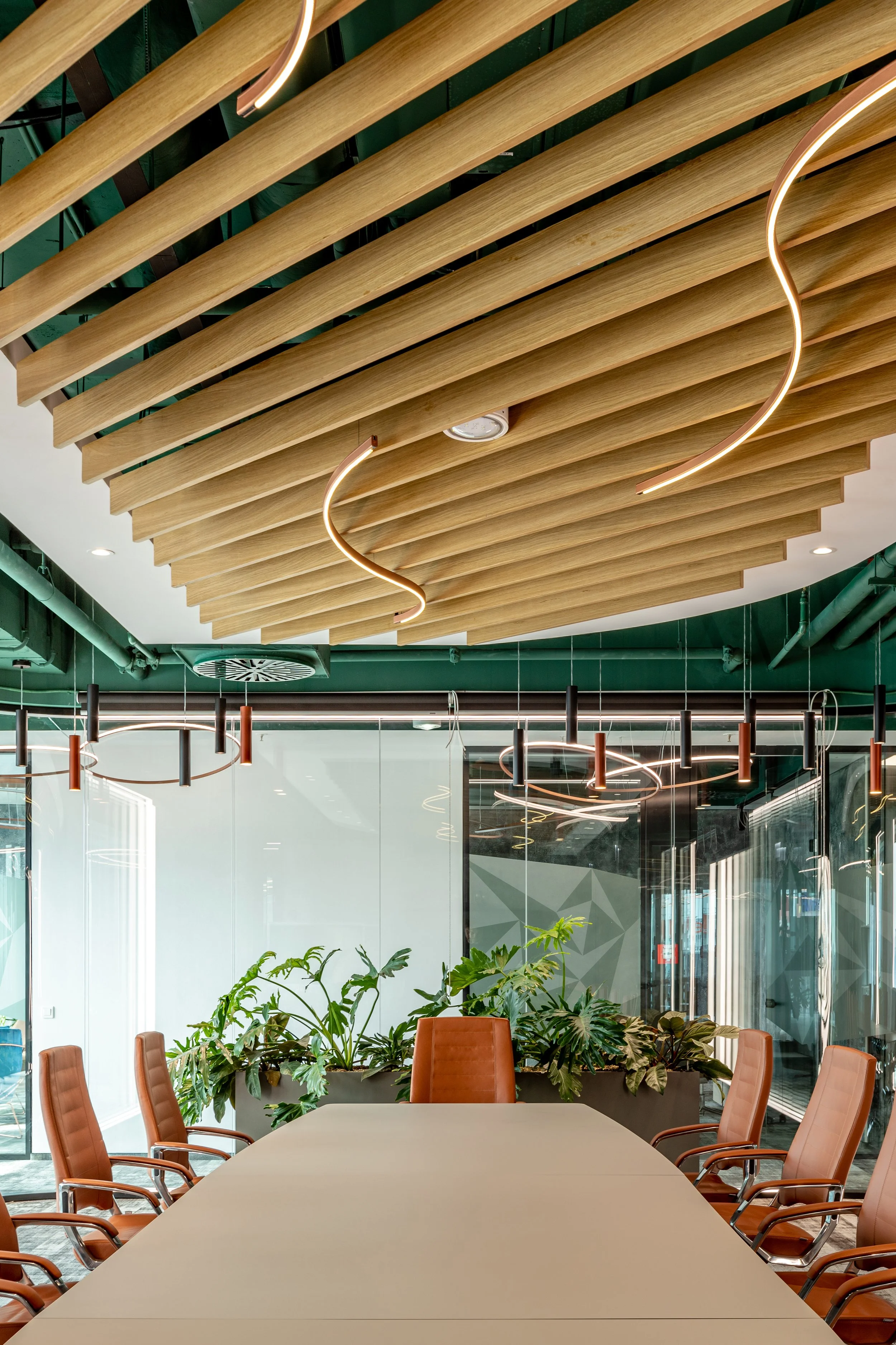 Modern conference room with a long table, brown leather chairs, indoor plants, glass wall, and decorative ceiling lighting fixtures.