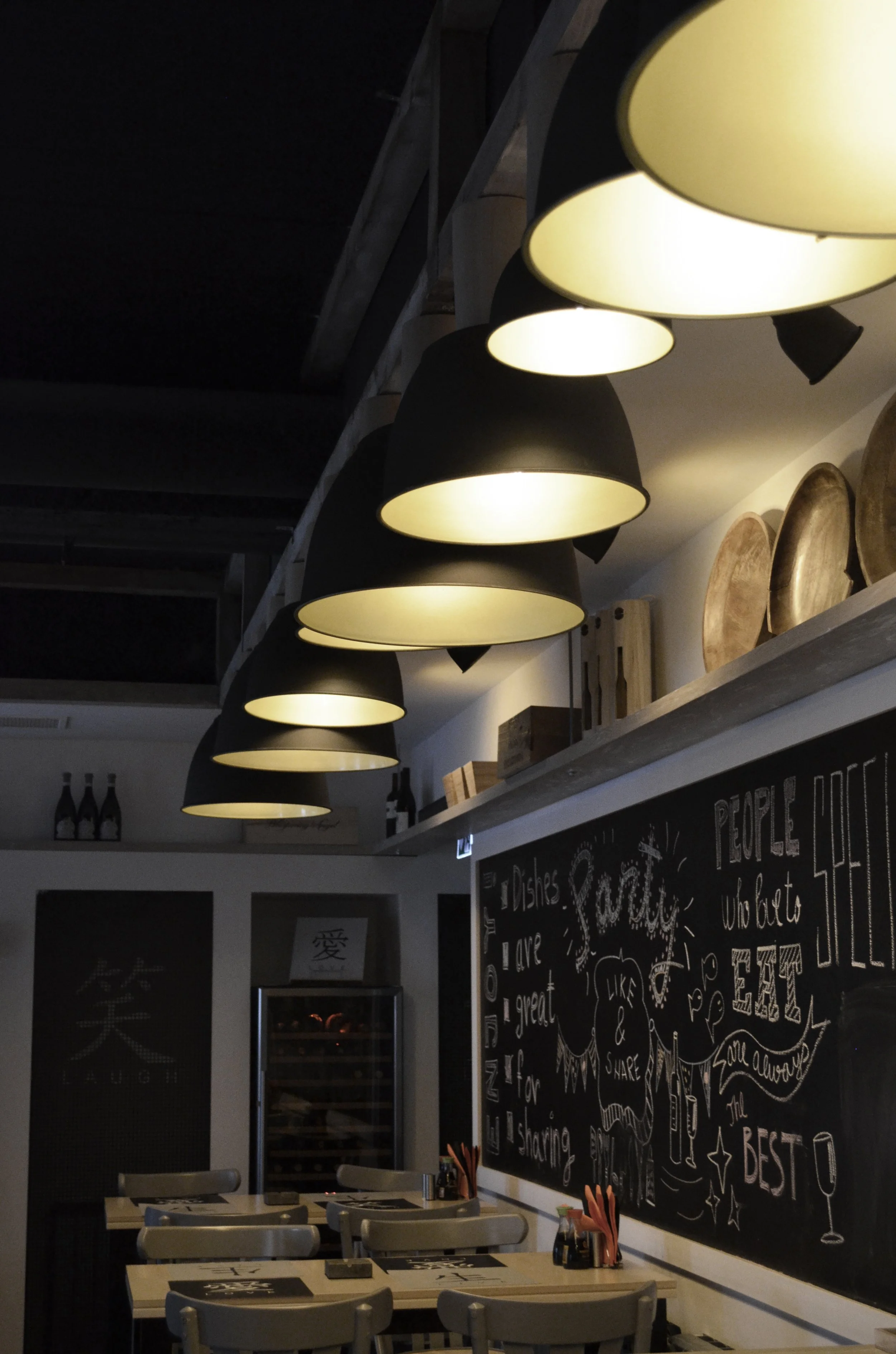 Interior of a restaurant with black pendant lights hanging from the ceiling, a chalkboard with decorative writing and drawings, tables set with menus and utensils, and a wine cooler in the background.