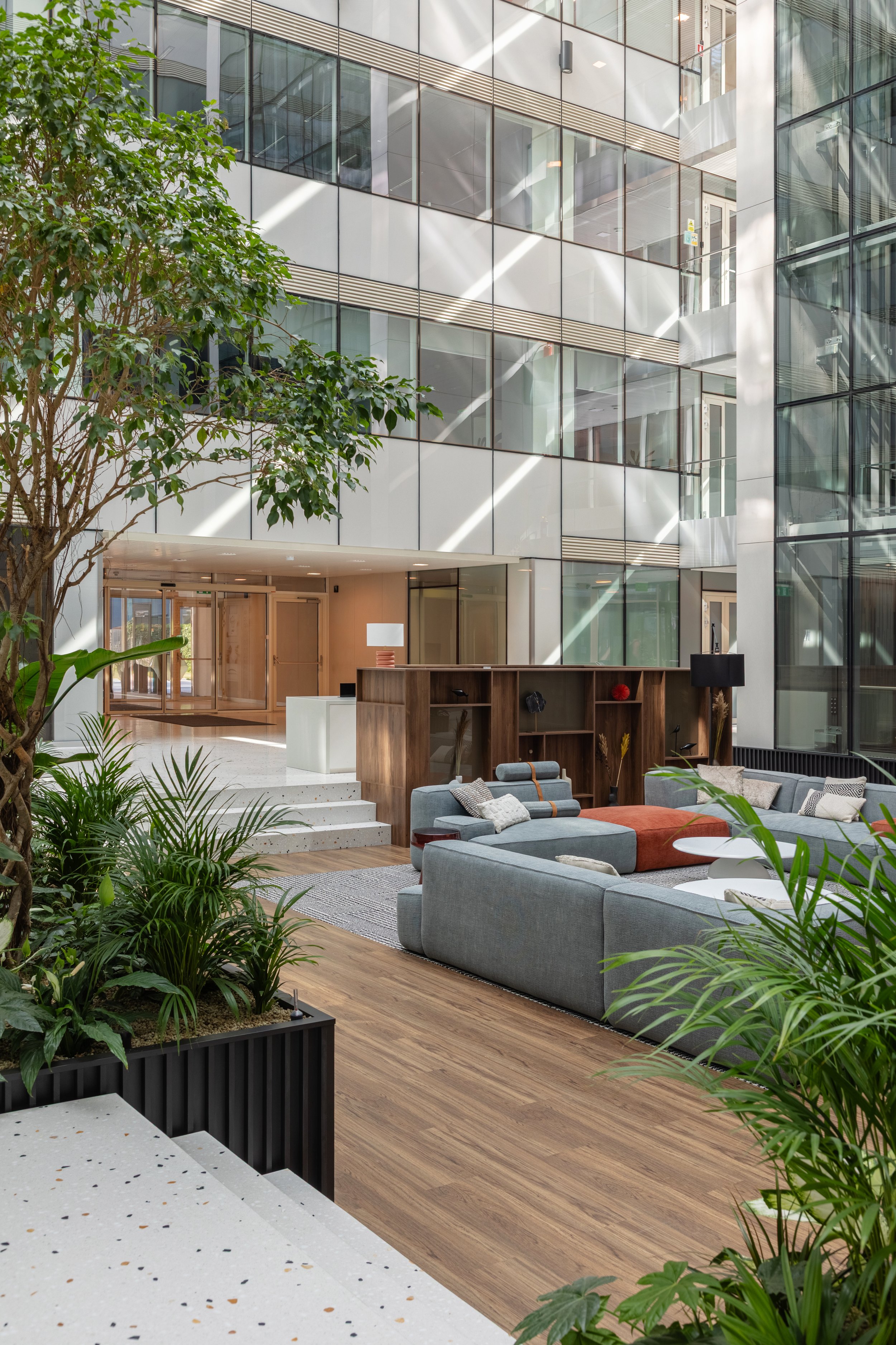 Interior view of a modern office lobby with large glass windows, green plants, and a seating area with gray sofas, a wooden bookshelf, and small white tables.