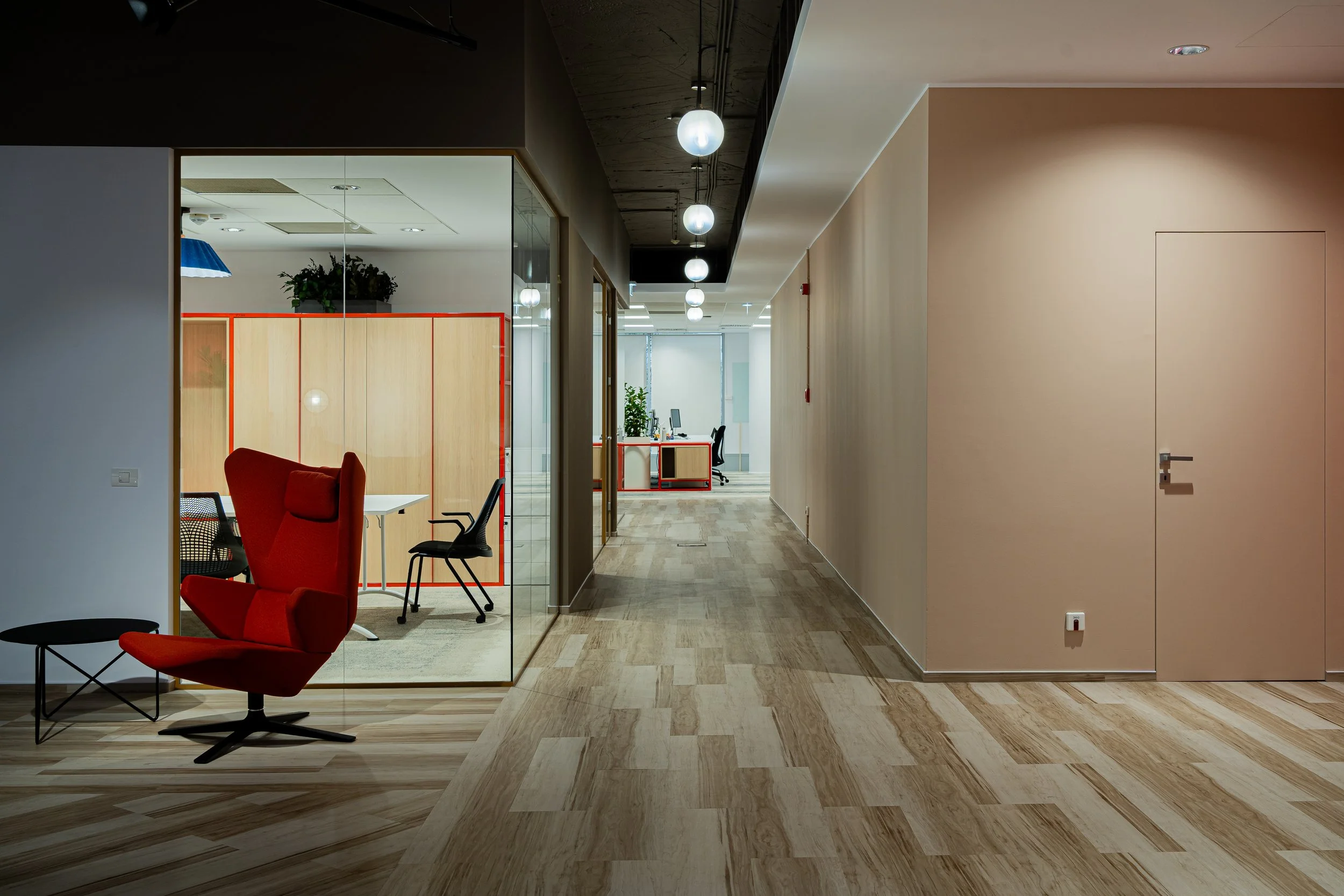 Modern office hallway with wooden floors, beige walls, and suspended ceiling lights. Conference room with glass walls on the left, featuring a red high-back chair, black chairs, and light wood furniture. Additional office space with desks and chairs visible further down the hallway.